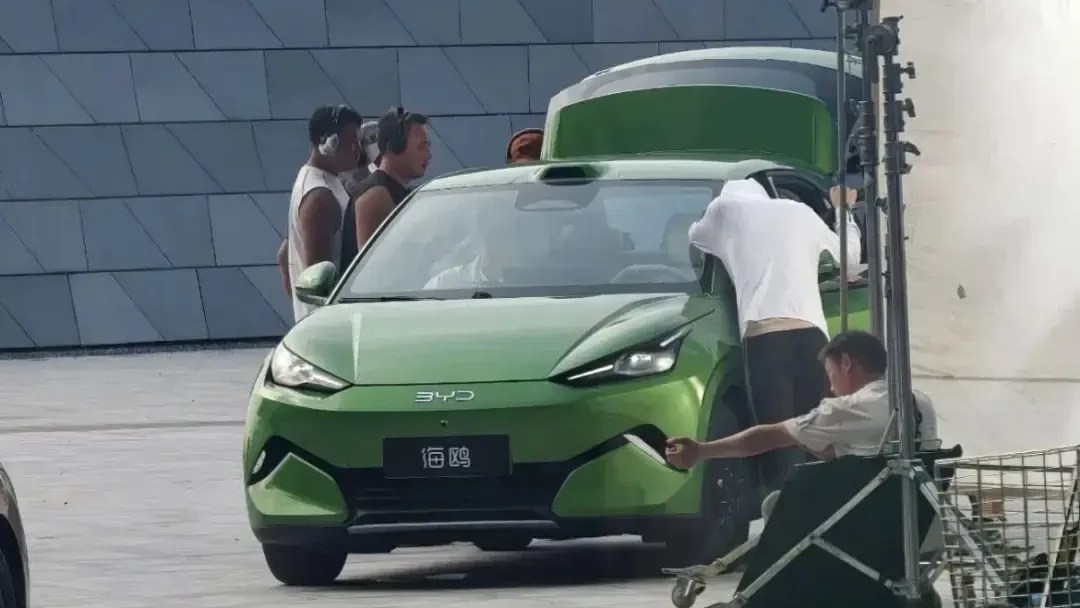 Carro verde elétrico BYD Seagull em gravação, com equipe de produção ao redor, incluindo um homem de camiseta branca ajustando algo no para-brisa e outro sentado em um carrinho de filmagem à direita