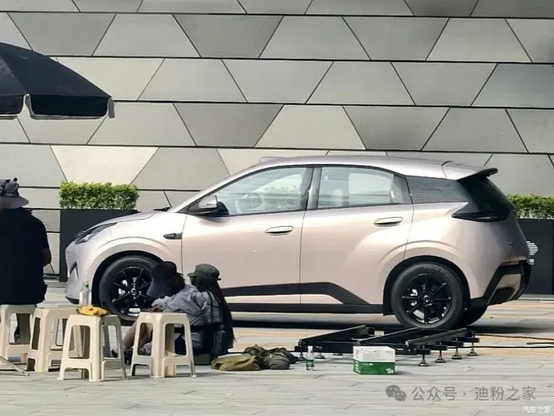 Carro hatchback cinza-claro com detalhes pretos, estacionado em frente a uma parede geométrica. Duas pessoas agachadas e uma sentada em bancos plásticos brancos, com equipamentos de filmagem no chão, sugerindo uma sessão de fotos ou gravação. Um guarda-sol preto aparece à esquerda