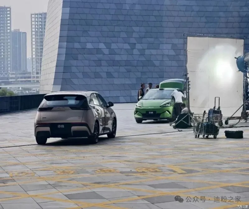Dois carros elétricos compactos em um set de filmagem externo. Um carro cinza-claro está em primeiro plano, visto por trás. Ao fundo, um carro verde-limão é ajustado por uma equipe de filmagem, com um grande refletor branco e equipamentos de iluminação próximos. Edifícios modernos e altos aparecem ao fundo, sob um céu nublado