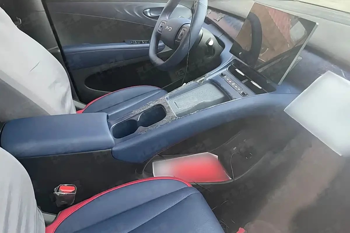 Interior de carro moderno com bancos azuis e detalhes vermelhos, console central com porta-copos e apoio de braço, volante multifuncional e tela multimídia grande no painel