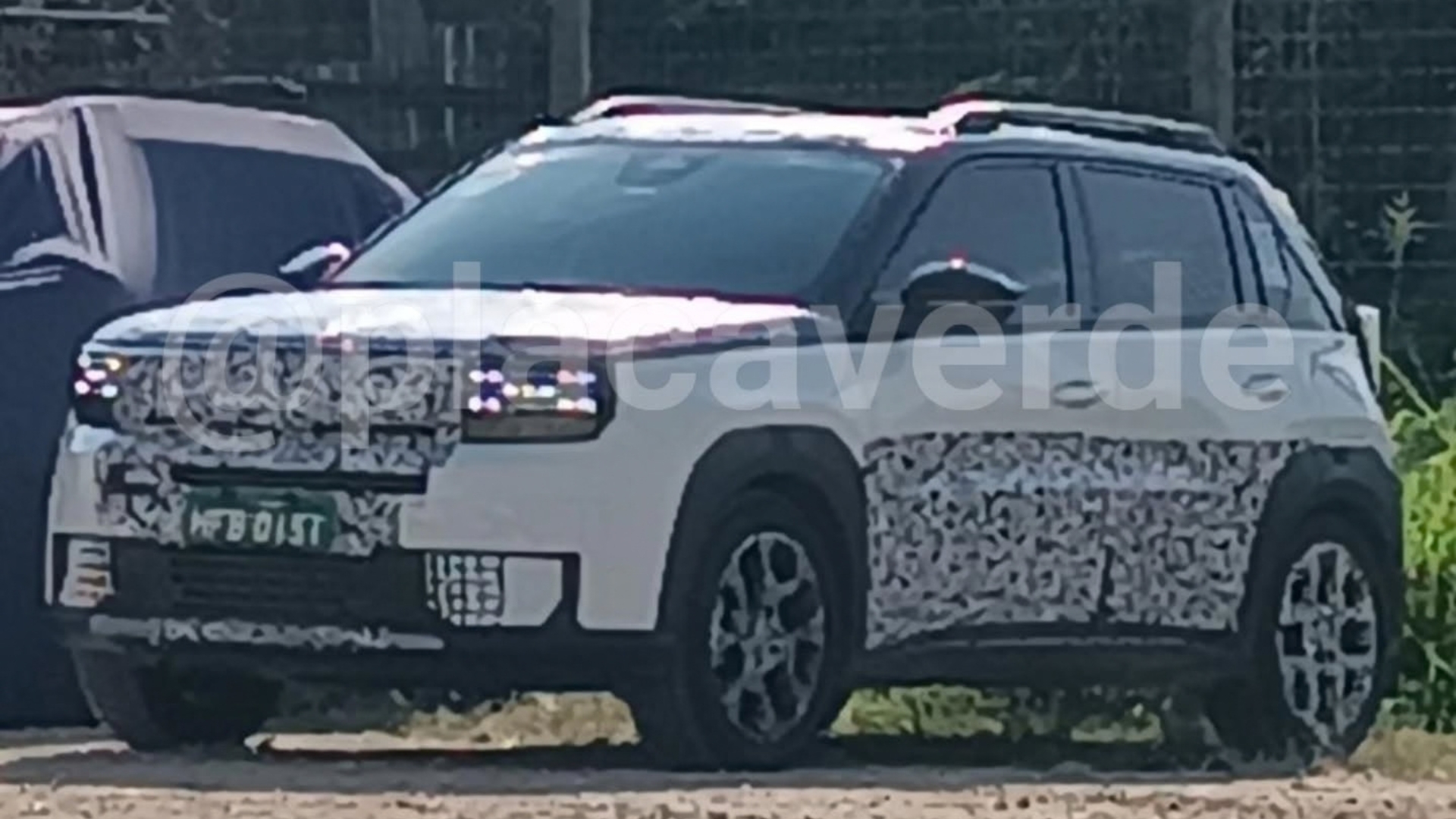 SUV branco camuflado com adesivos cinzas e pretos, teto preto, rack de teto, faróis acesos e placa verde HFB 01ST, estacionado em terreno de terra com outro veículo escuro ao fundo