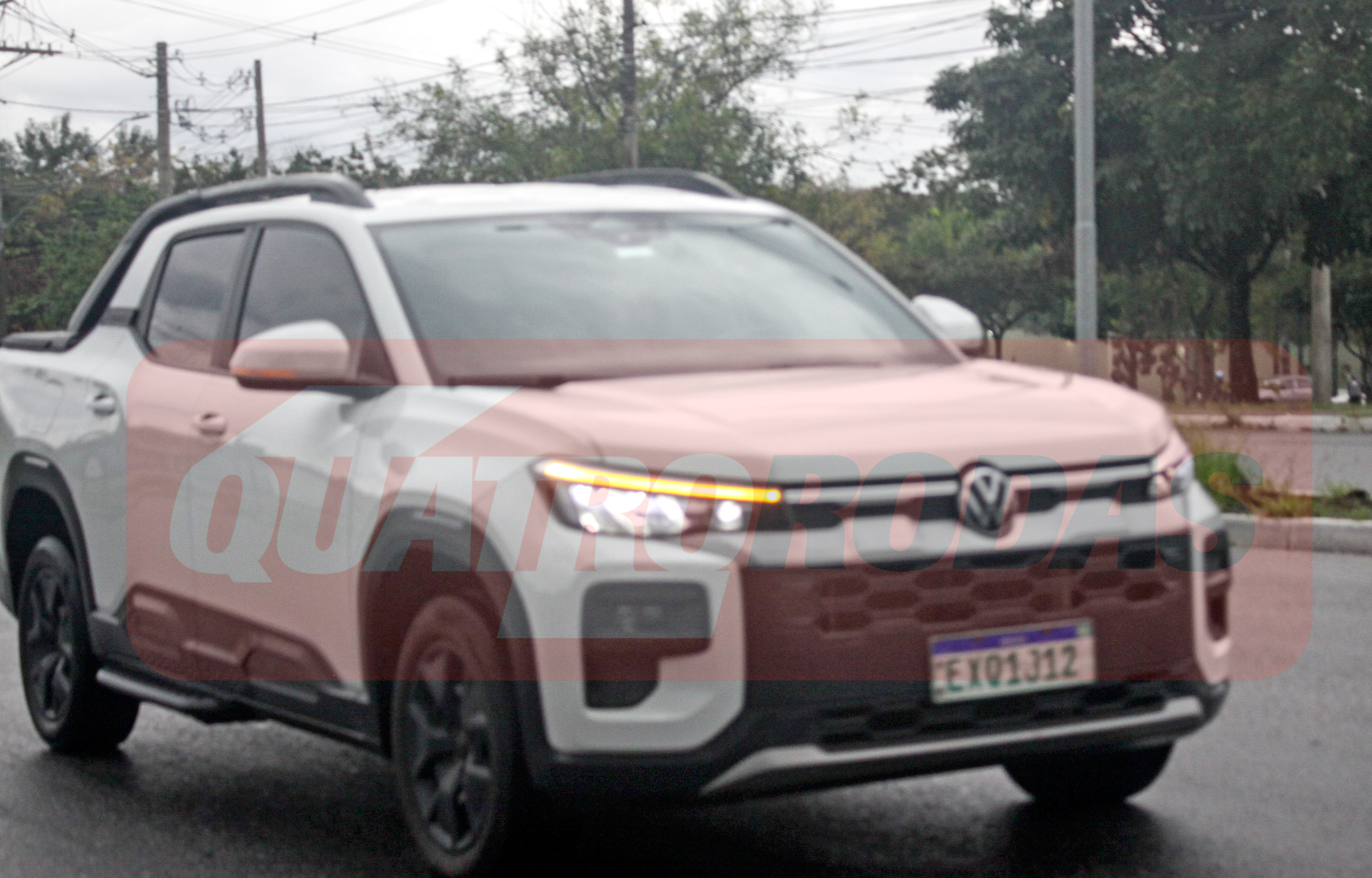 Picape branca com detalhes em preto e teto solar, rodando em rua asfaltada com árvores ao fundo. A frente do veículo tem faróis acesos e grade escura com o logo da Volkswagen. A placa de teste é EXQ1J12