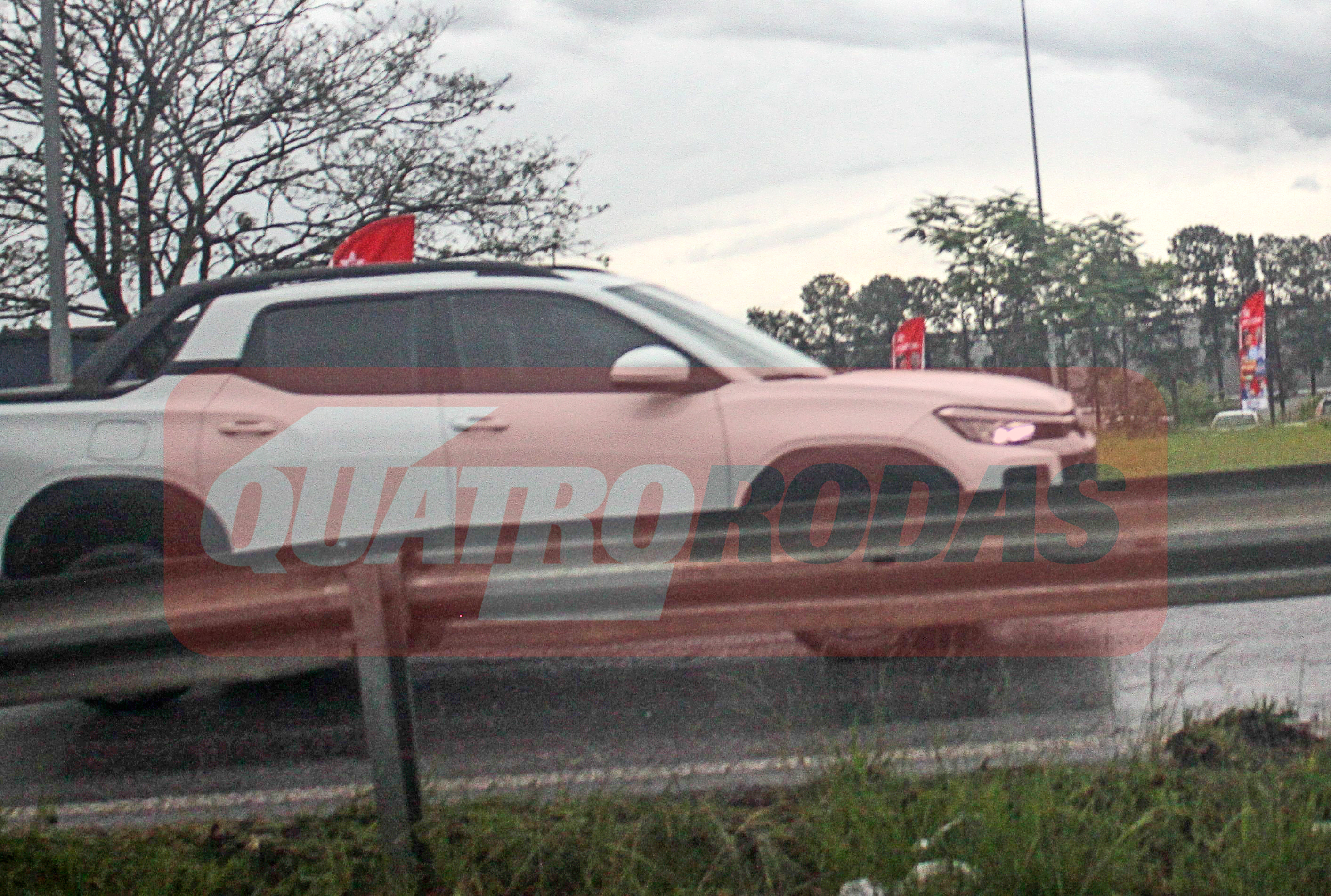 Picape branca camuflada com adesivos rosa e um pequeno sinal vermelho no teto, trafegando em uma estrada com guard-rail cinza. O céu está nublado e há árvores ao fundo. A marca dágua QUATRO RODAS está sobreposta na imagem
