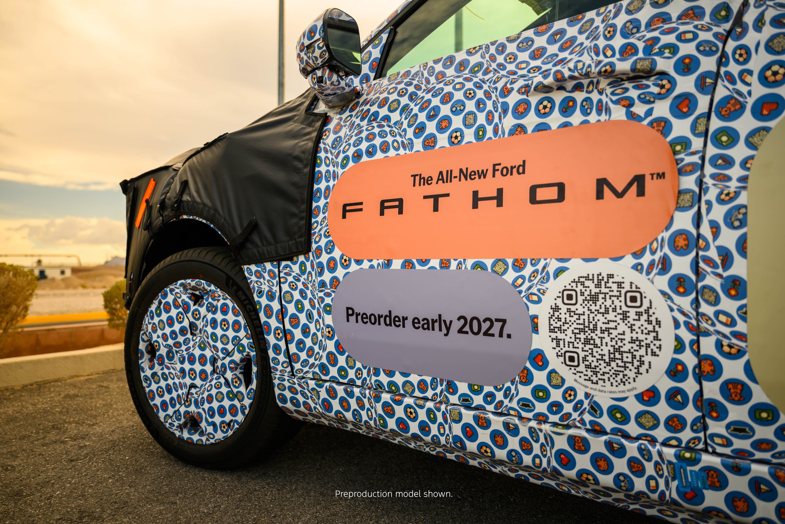 Lateral de um carro protótipo Ford Fathom com camuflagem azul e branca, adesivos laranja e cinza com Preorder early 2027 e um QR code. O para-lama dianteiro está coberto por tecido preto, e o céu laranja do pôr do sol aparece ao fundo