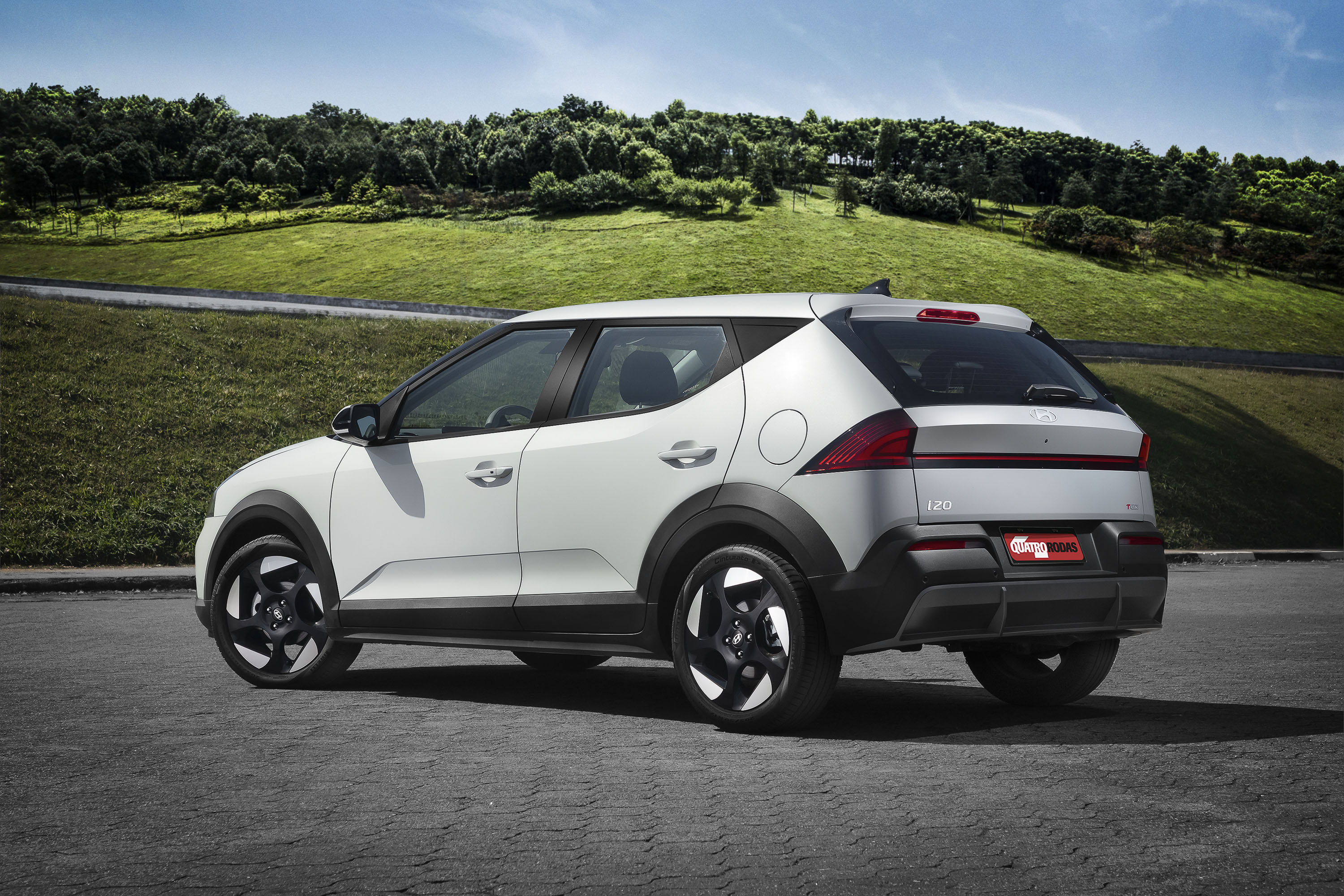 Carro branco com detalhes pretos, modelo SUV compacto, estacionado em asfalto cinza, com uma colina verde e árvores ao fundo sob céu azul