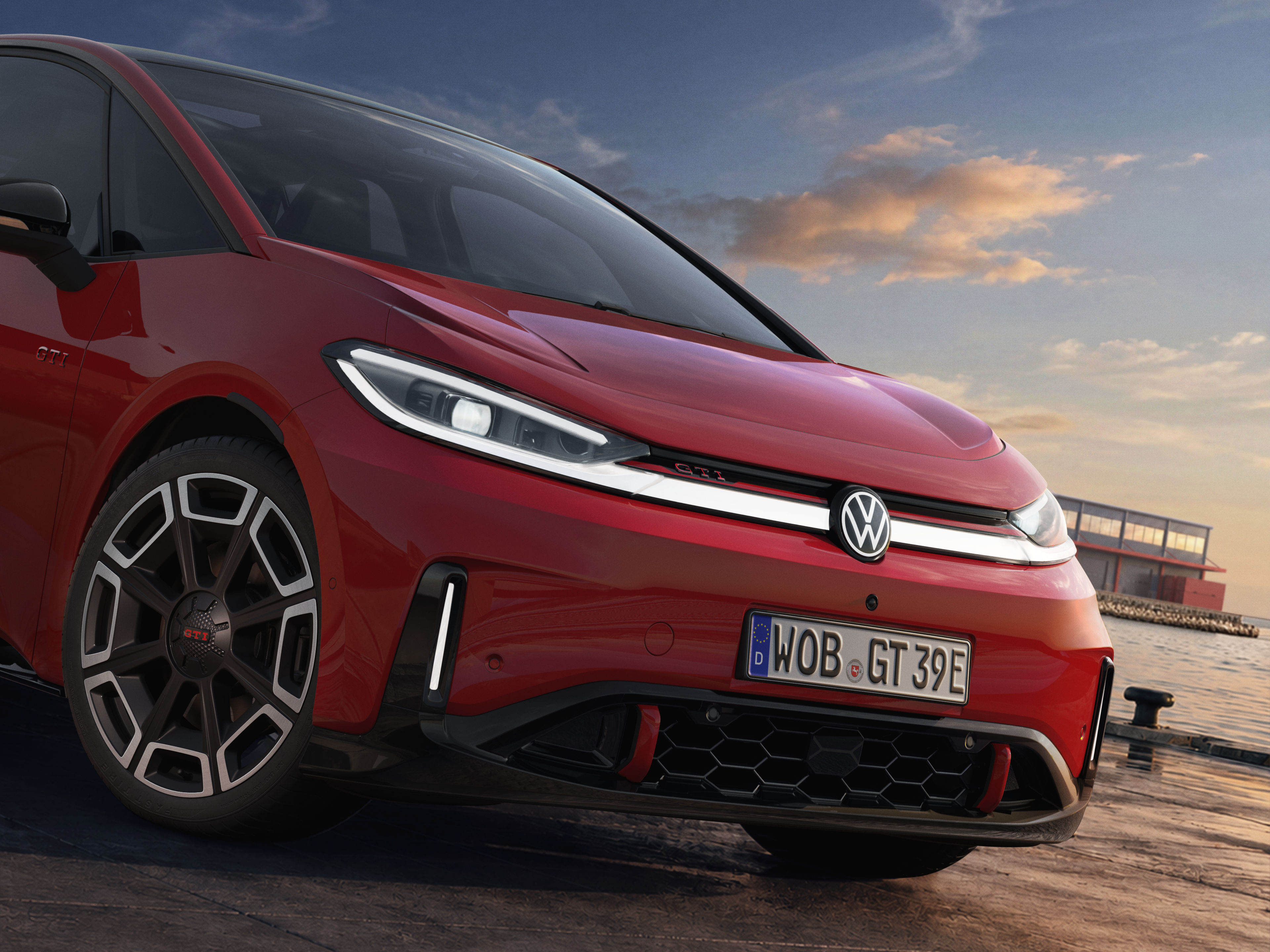 Carro elétrico Volkswagen ID. GTI vermelho, com faróis de LED acesos, grade frontal preta e rodas esportivas, estacionado em um píer ao entardecer, com o mar e um edifício ao fundo