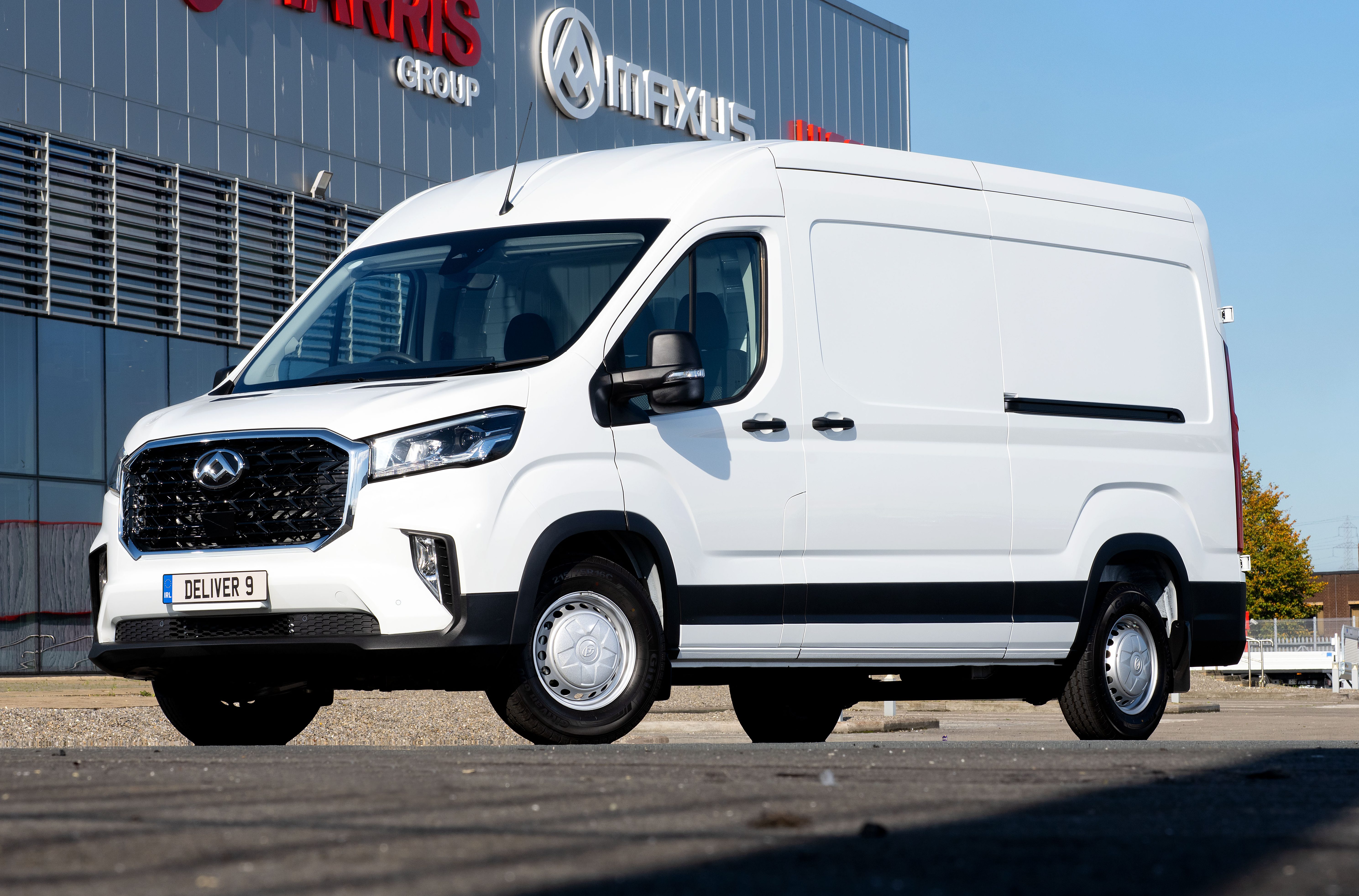 Furgão branco Maxus Deliver 9 estacionado em frente a um prédio cinza com os logotipos Harris Group e Maxus em vermelho e prata, sob um céu azul claro