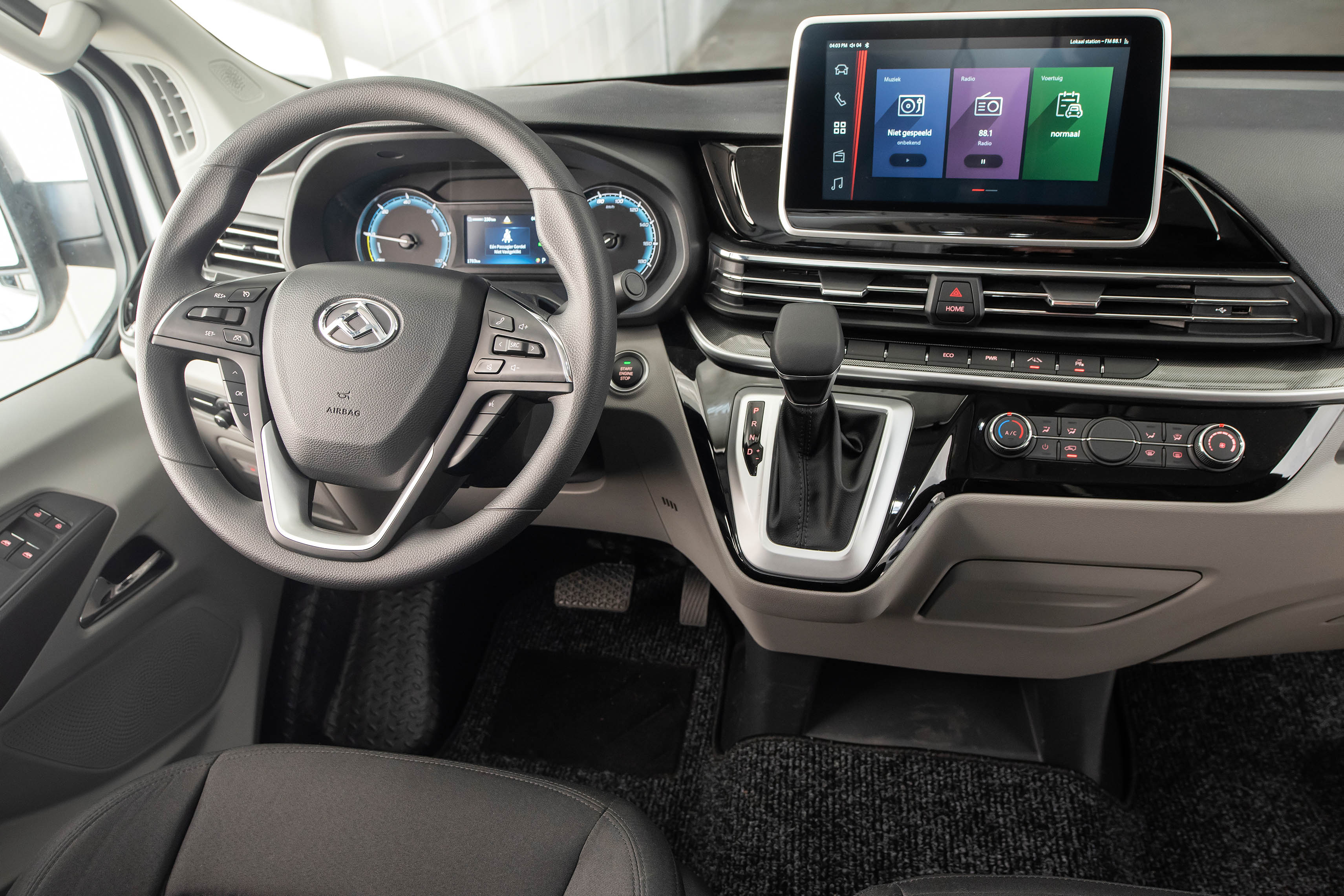 Interior de um carro moderno, com volante multifuncional preto e cinza, painel digital azul, tela multimídia sensível ao toque exibindo ícones de aplicativos, câmbio automático e controles de ar-condicionado. O ambiente é predominantemente cinza e preto, com detalhes cromados, transmitindo funcionalidade e tecnologia