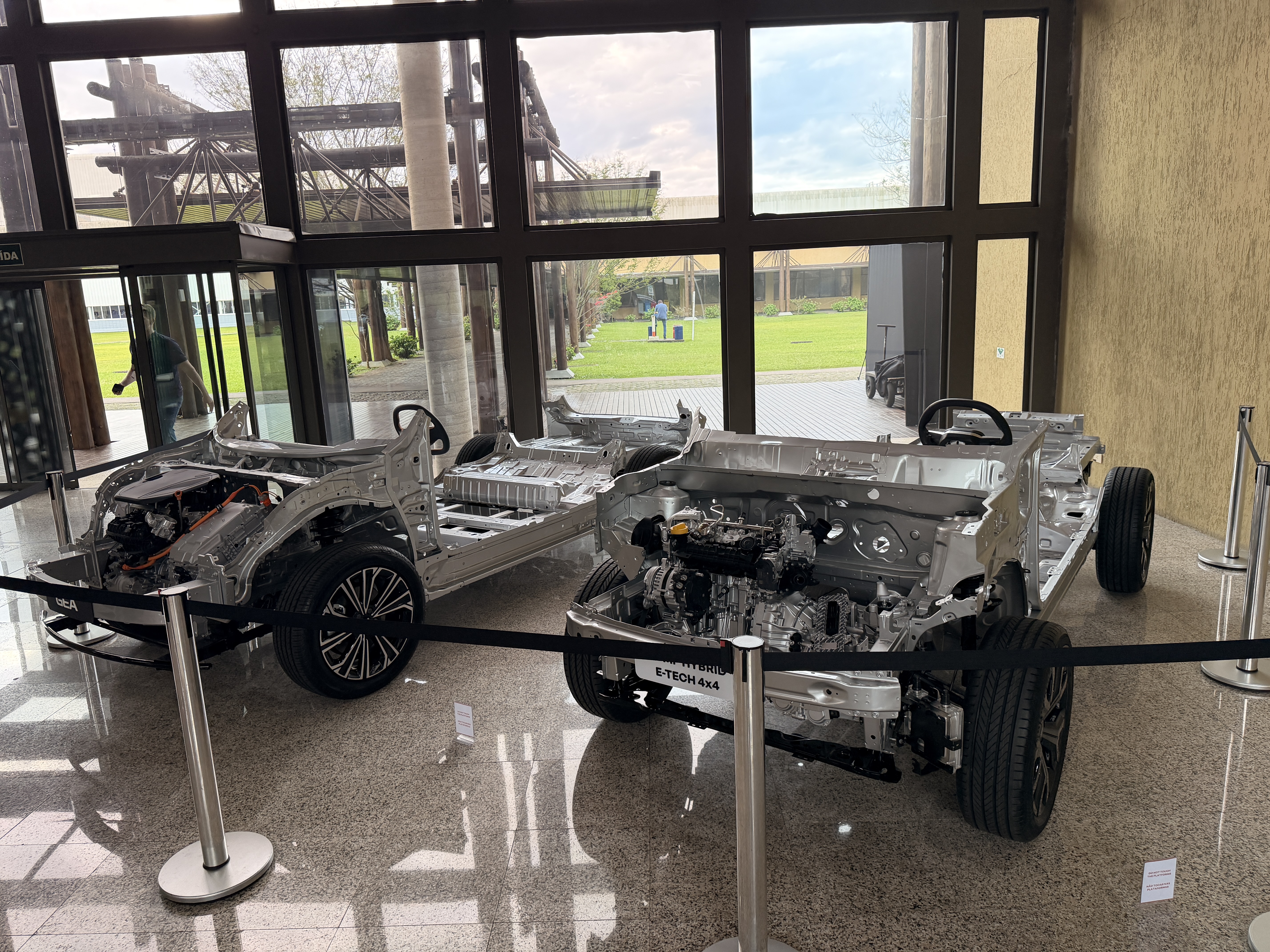 Dois chassis de carros expostos em um salão com grandes janelas. O chassi da frente exibe o motor e a suspensão, com a inscrição E-TECH 4X4. O chassi de trás mostra a estrutura sem motor. Ambos estão cercados por cordões de isolamento. Ao fundo, um gramado verde e árvores são visíveis através das janelas