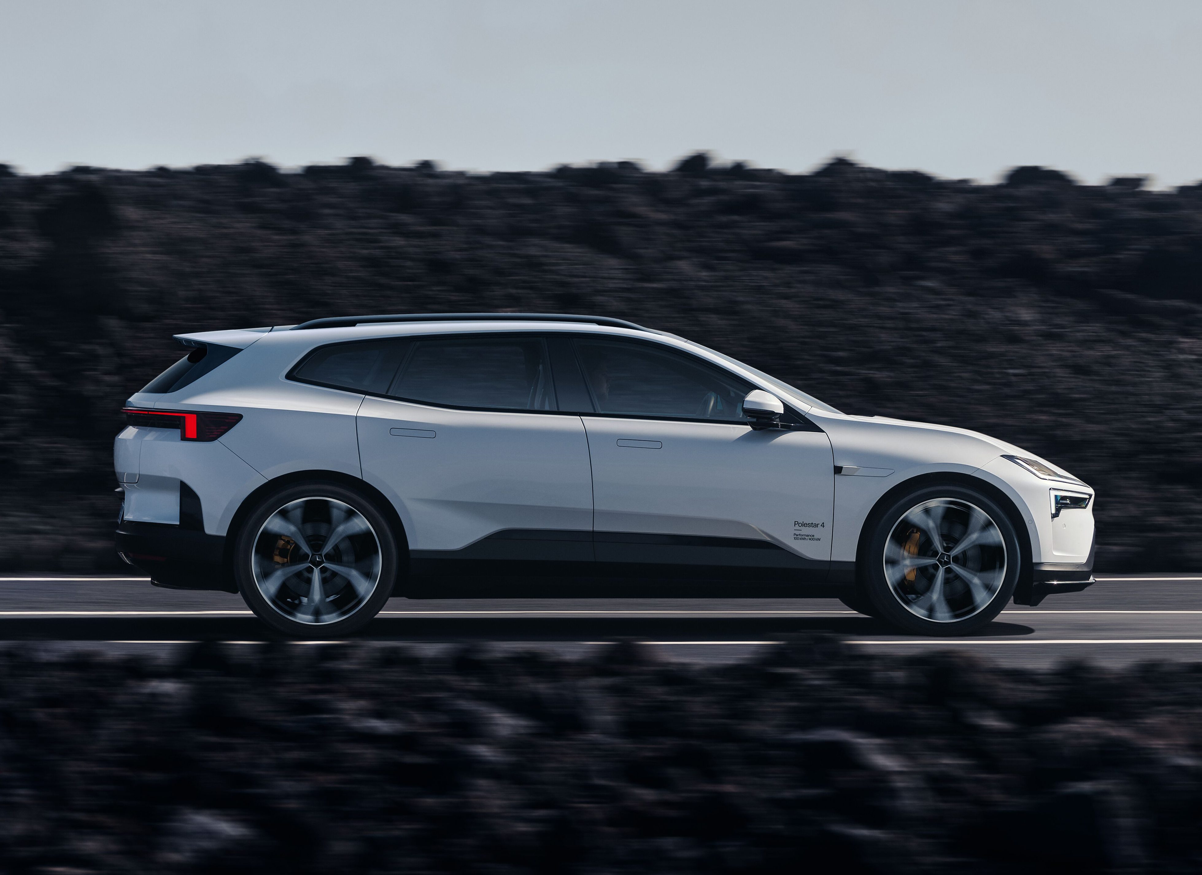 Um carro SUV branco moderno em movimento, visto de lado, em uma estrada asfaltada. O veículo tem rodas esportivas e detalhes escuros na parte inferior. Ao fundo, um terreno rochoso escuro e um céu claro.