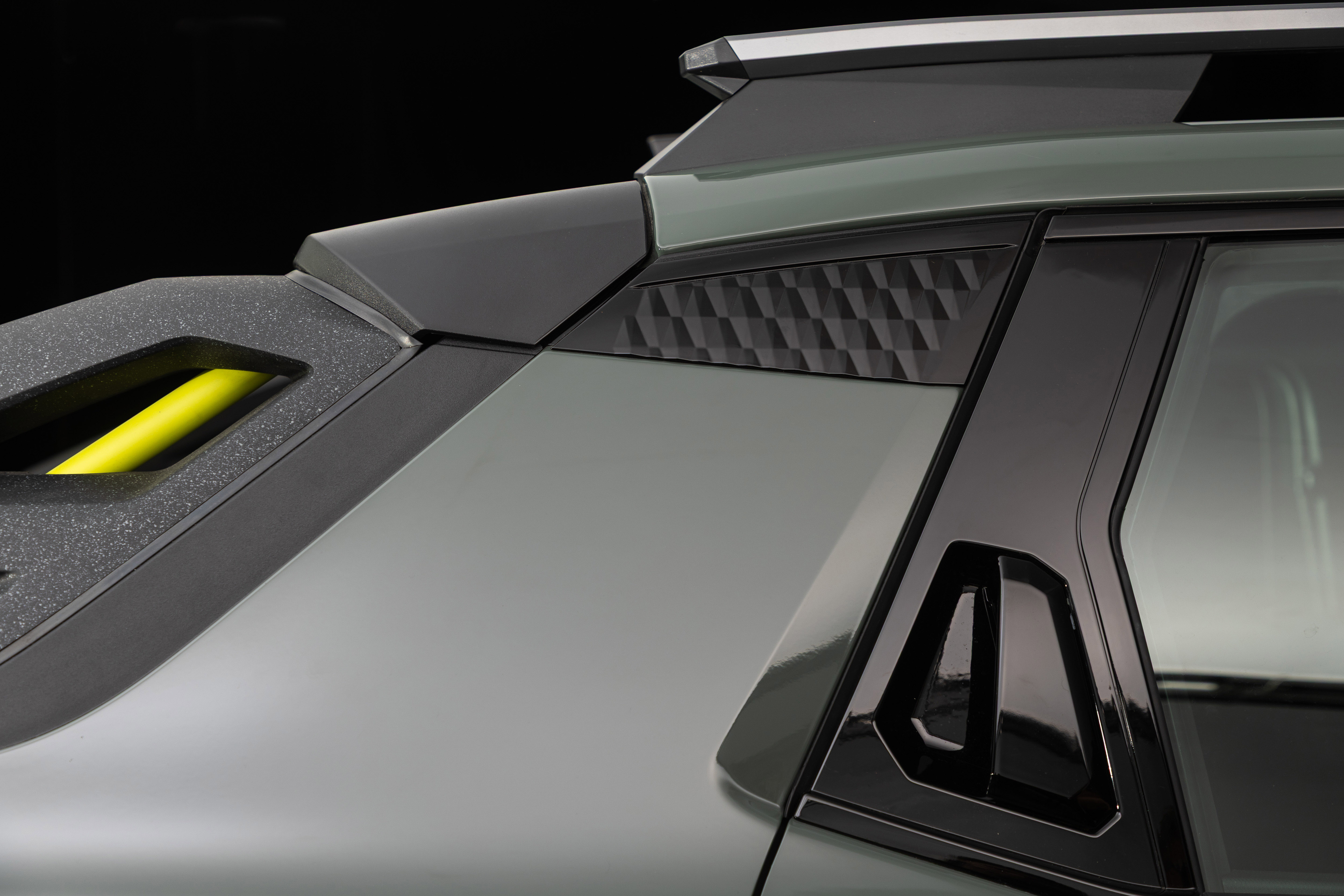Lateral de um carro verde-acinzentado com detalhes em preto e amarelo. O teto tem um rack prateado, e a coluna C exibe um padrão geométrico em relevo. Uma alça amarela se destaca na parte superior da porta traseira, que tem uma maçaneta preta integrada. O fundo é escuro