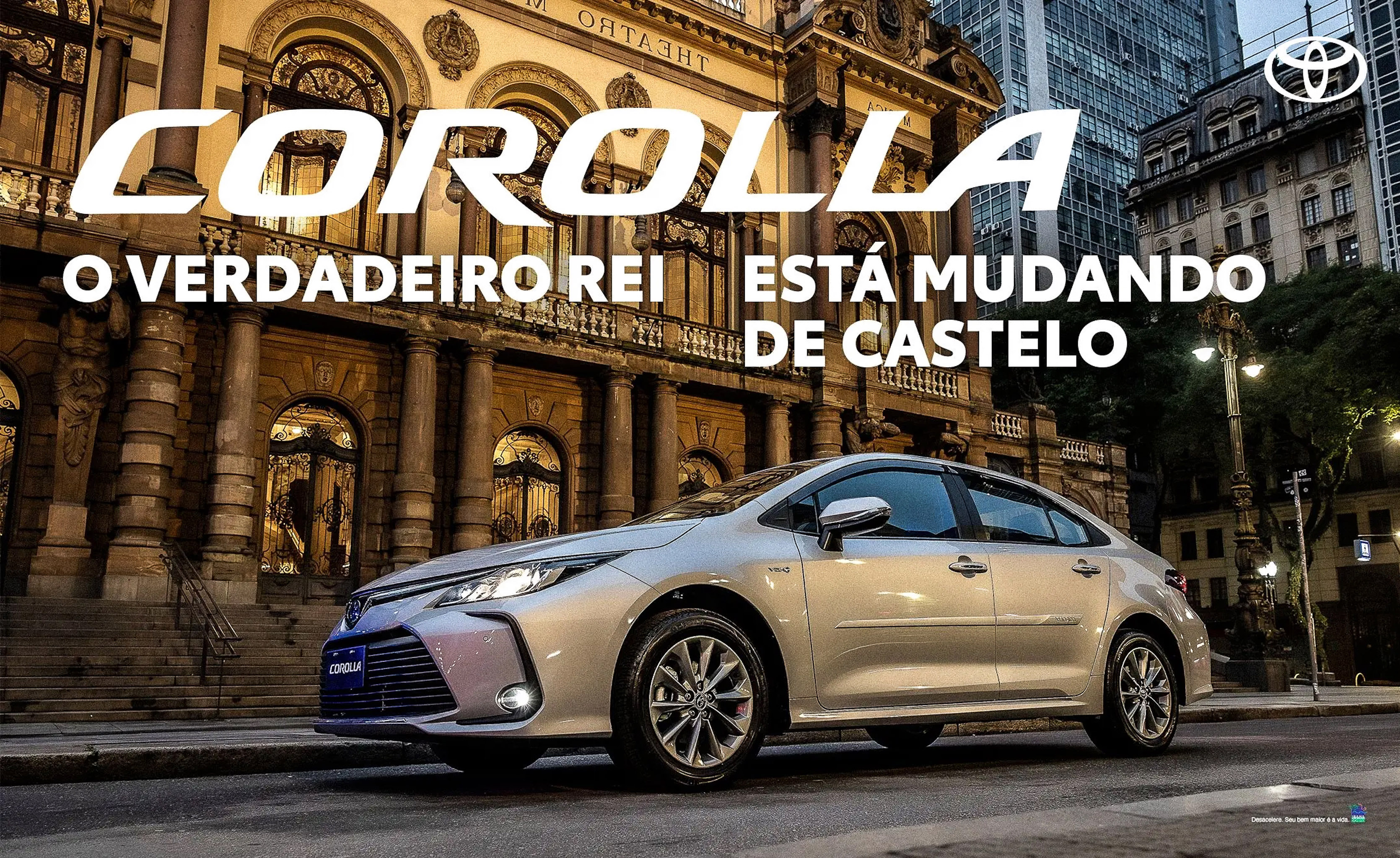 Carro Toyota Corolla prata estacionado em rua urbana à noite, com um edifício histórico iluminado ao fundo. O texto COROLLA O VERDADEIRO REI ESTÁ MUDANDO DE CASTELO aparece em destaque