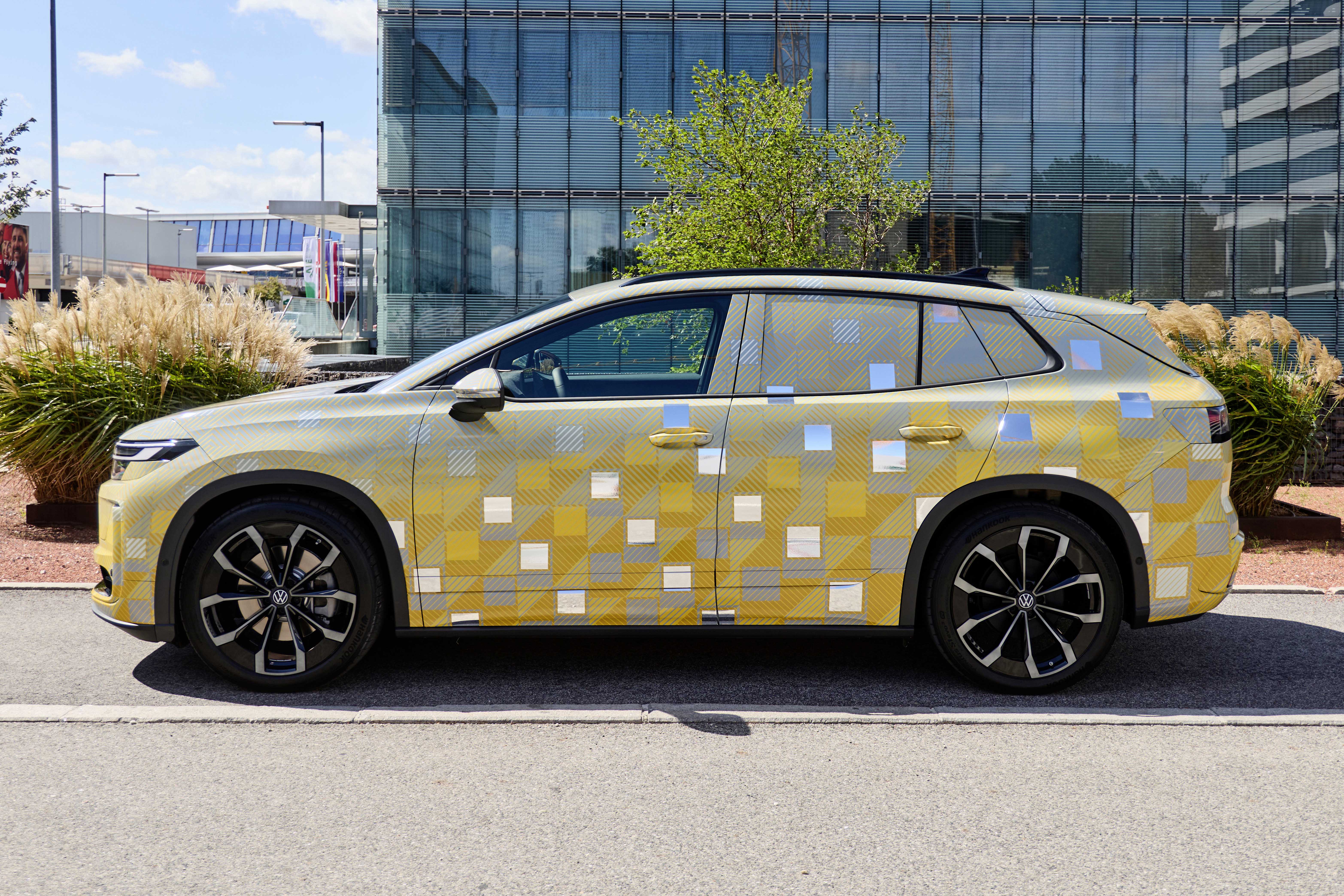 Carro camuflado em tons de amarelo, cinza e branco, com quadrados espelhados, estacionado em frente a um prédio de vidro e arbustos