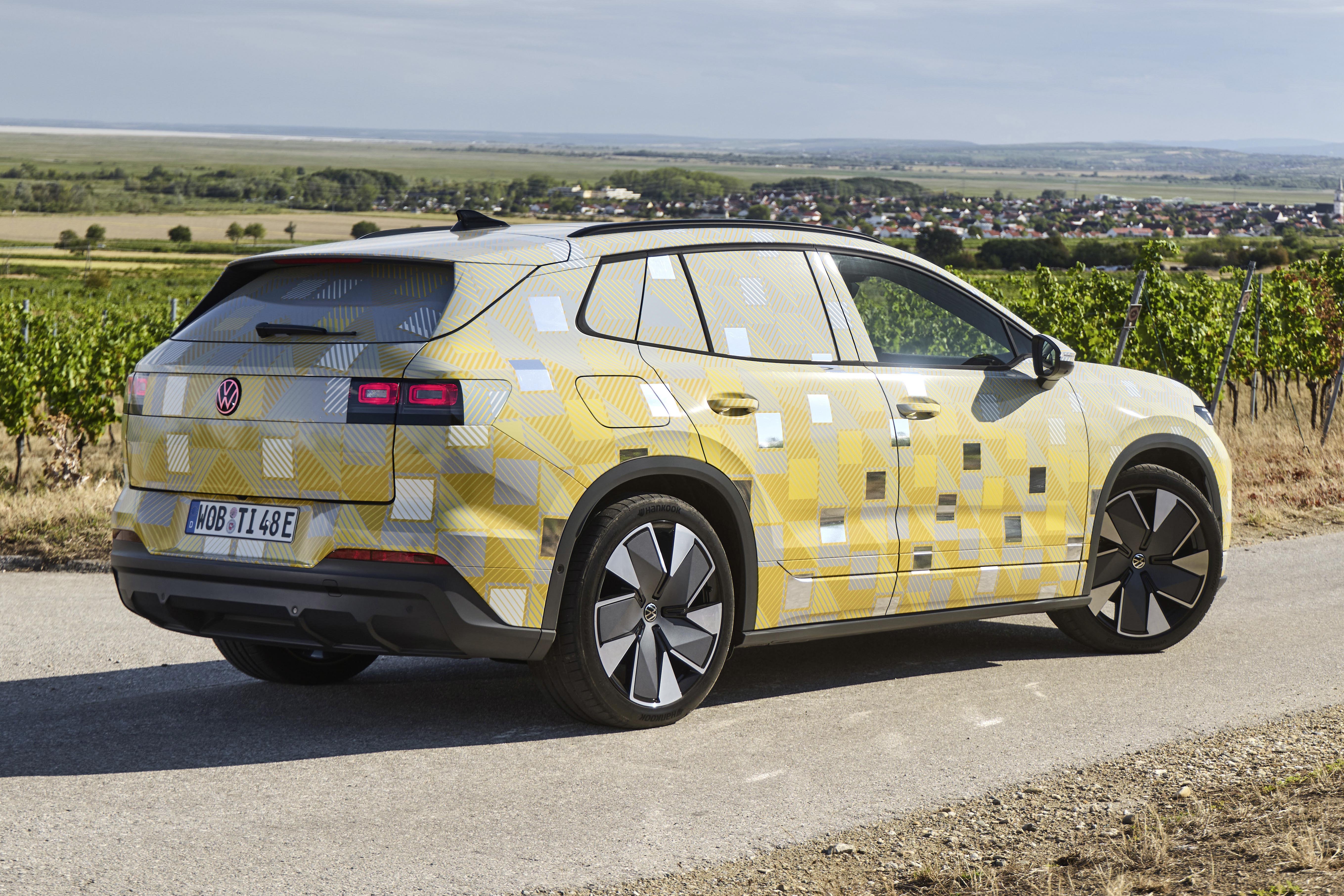 SUV Volkswagen camuflado com adesivos amarelos e cinzas, visto de traseira lateral, em estrada de terra com vinhedos e uma cidade ao fundo