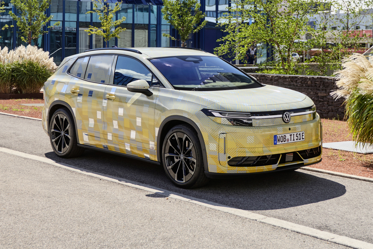 Carro SUV Volkswagen camuflado com adesivos amarelos e brancos, estacionado em uma rua asfaltada com meio-fio branco. Ao fundo, um prédio moderno de vidro e árvores verdes