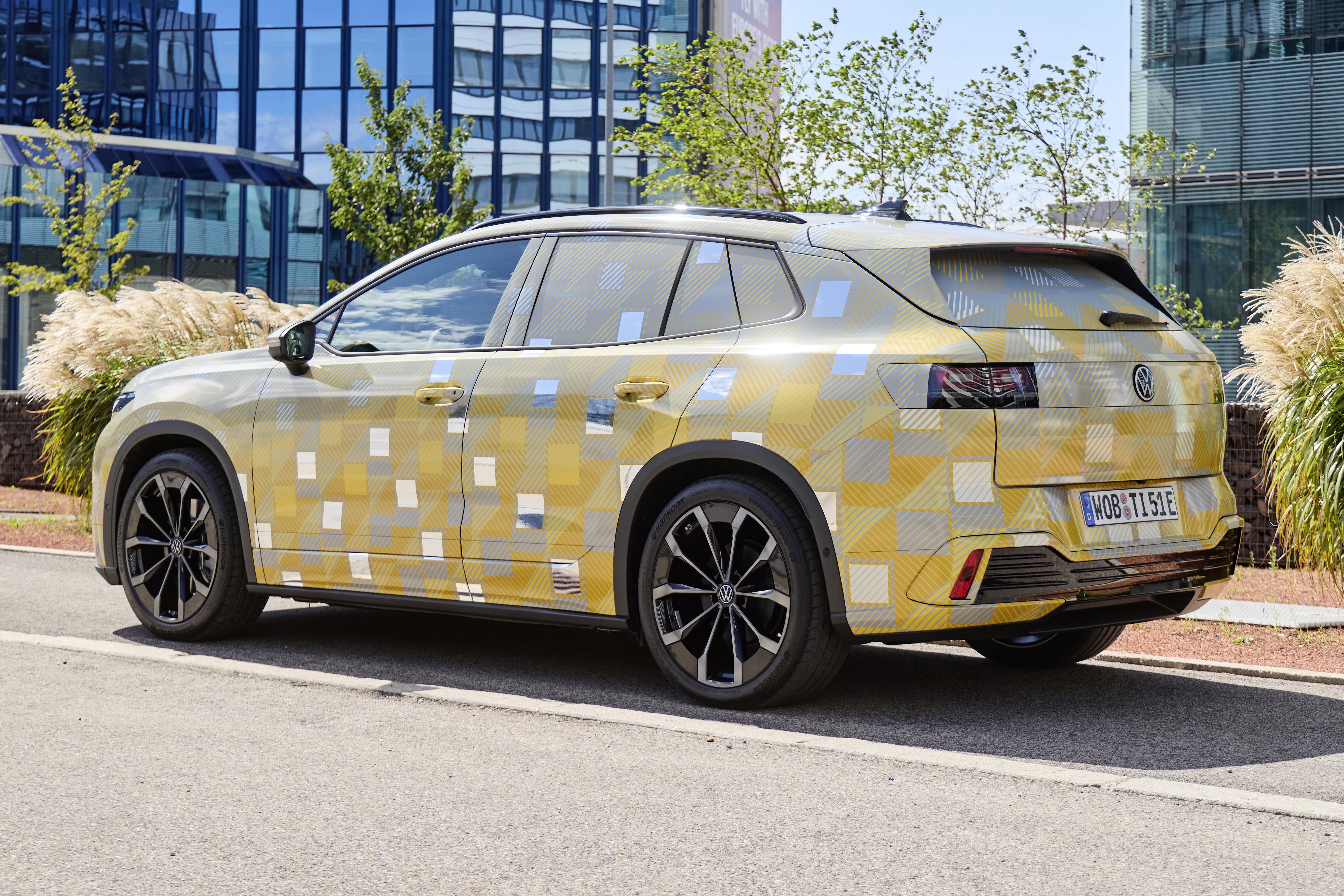SUV camuflado com adesivos amarelos, cinzas e quadrados prateados, estacionado em rua asfaltada. Ao fundo, prédios de vidro azul refletem o céu e árvores verdes.