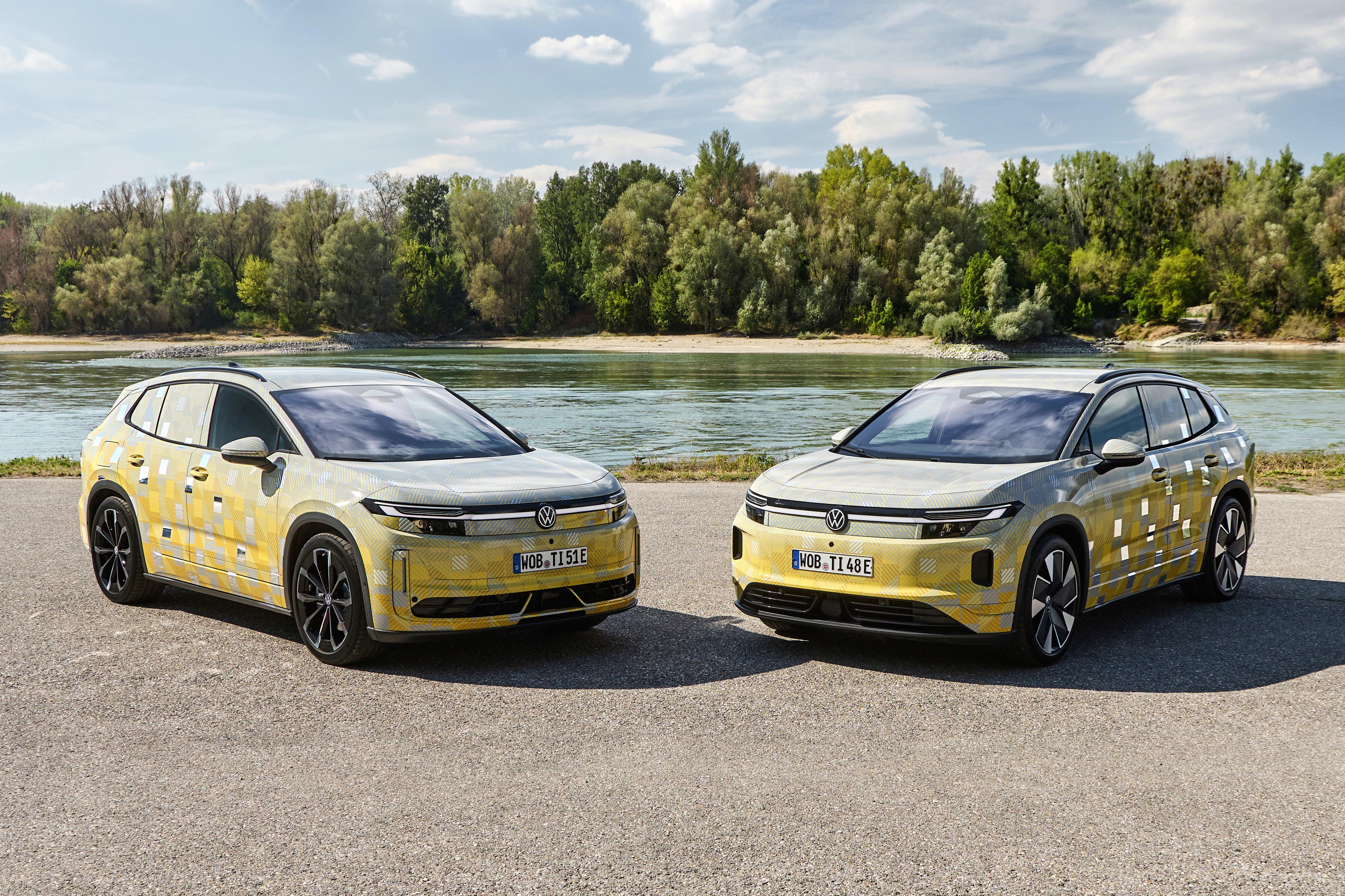 Dois carros Volkswagen elétricos, camuflados em amarelo e cinza, estacionados lado a lado em uma estrada de asfalto. Ao fundo, um rio e uma floresta densa sob céu azul com nuvens esparsas