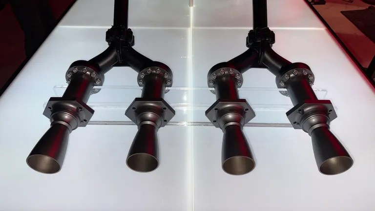 Duas estruturas metálicas escuras, cada uma com duas saídas em forma de cone, dispostas simetricamente sobre uma superfície branca iluminada
