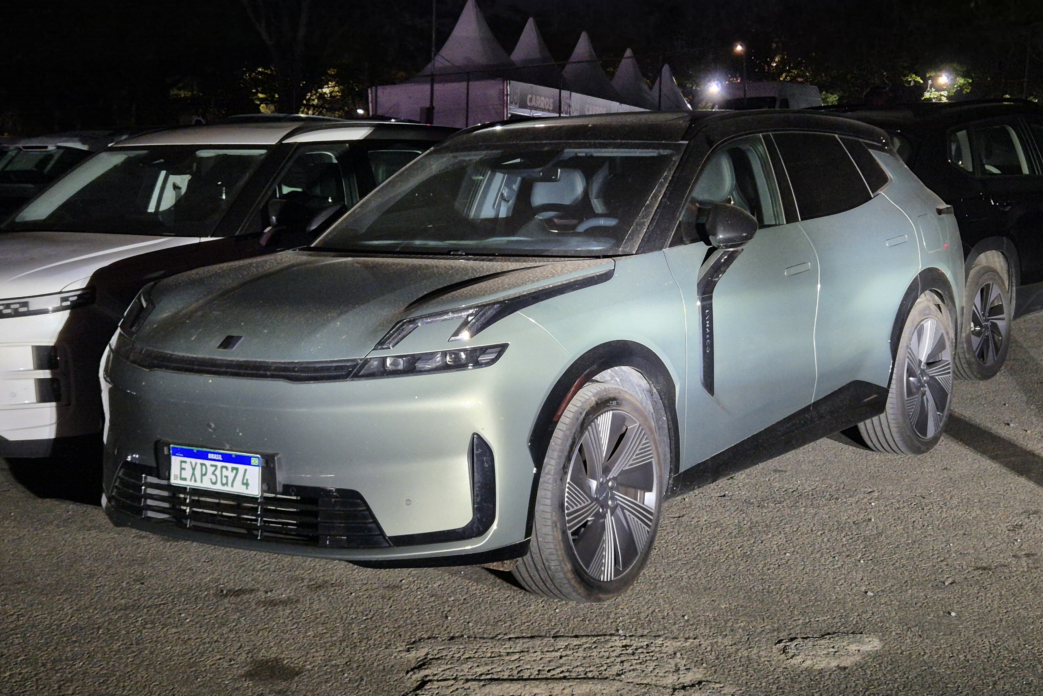Carro elétrico cinza-esverdeado, com design futurista e placa brasileira EXP3G74, estacionado à noite. Ao fundo, outros carros e tendas brancas iluminadas
