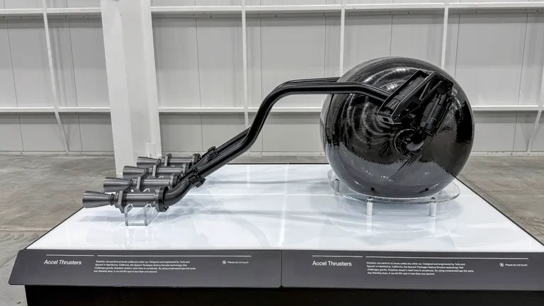 Um sistema de propulsão com um tanque esférico preto brilhante e seis propulsores cinza metálicos conectados por tubos pretos, exibido em uma plataforma branca com texto Accel Thrusters e instruções