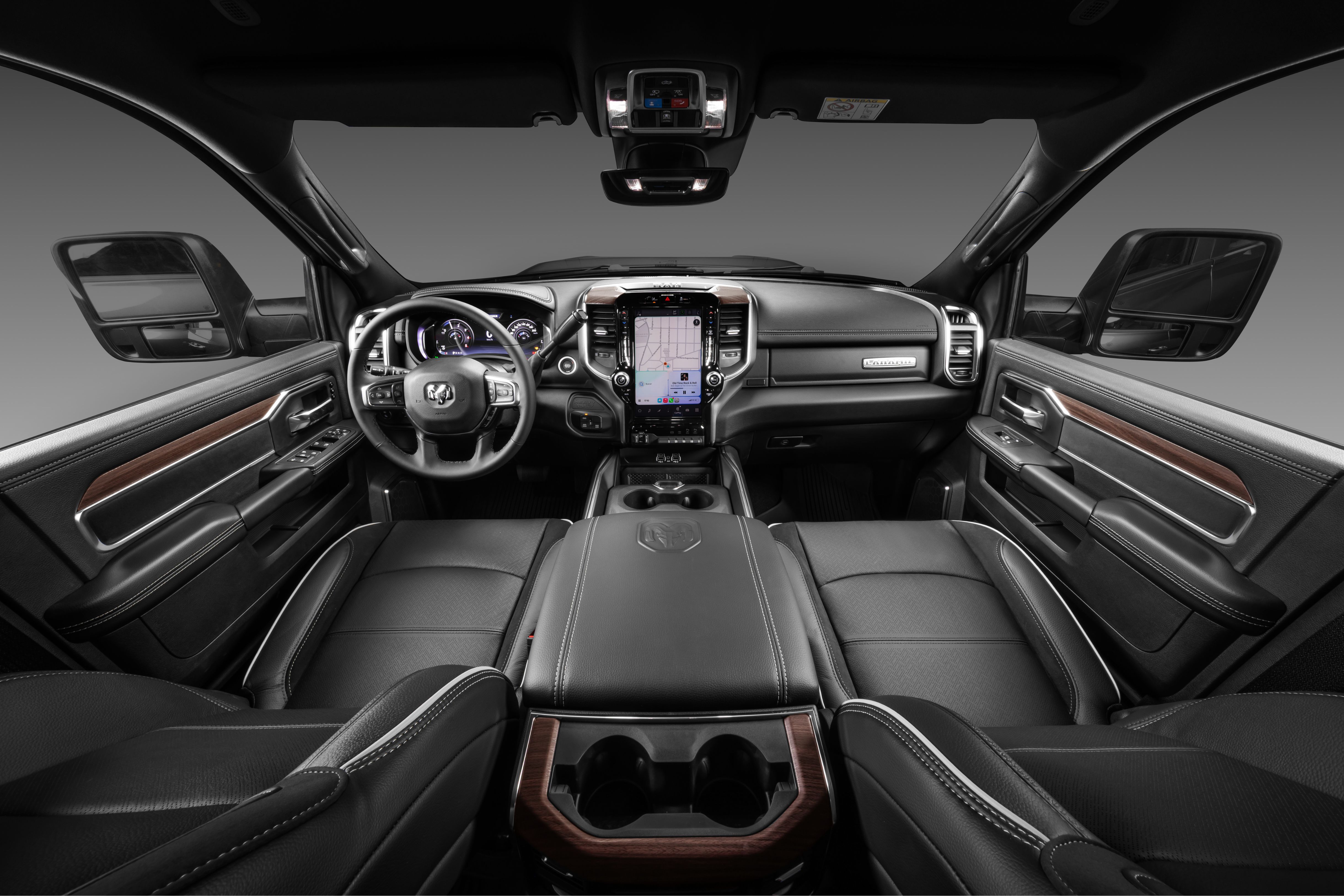 Interior de uma picape Ram, com bancos de couro preto e costura branca. O painel tem uma tela central grande exibindo um mapa, volante multifuncional e detalhes em madeira escura nas portas e console central. O ambiente é sofisticado e moderno