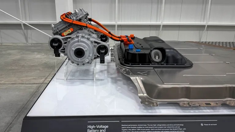 Um motor elétrico cinza com cabos laranja e uma bateria de carro prateada em exposição sobre uma plataforma branca