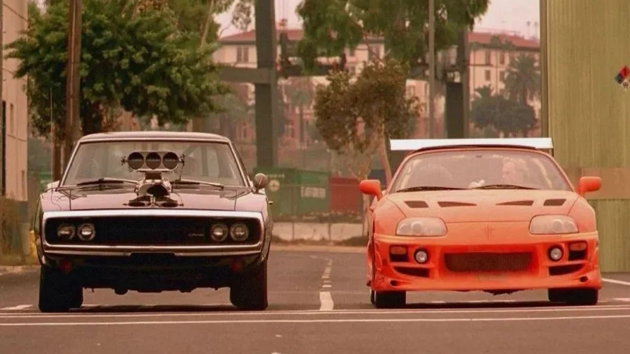 Dois carros de corrida, um Dodge Charger preto com motor exposto e um Toyota Supra laranja com aerofólio, lado a lado em uma rua urbana