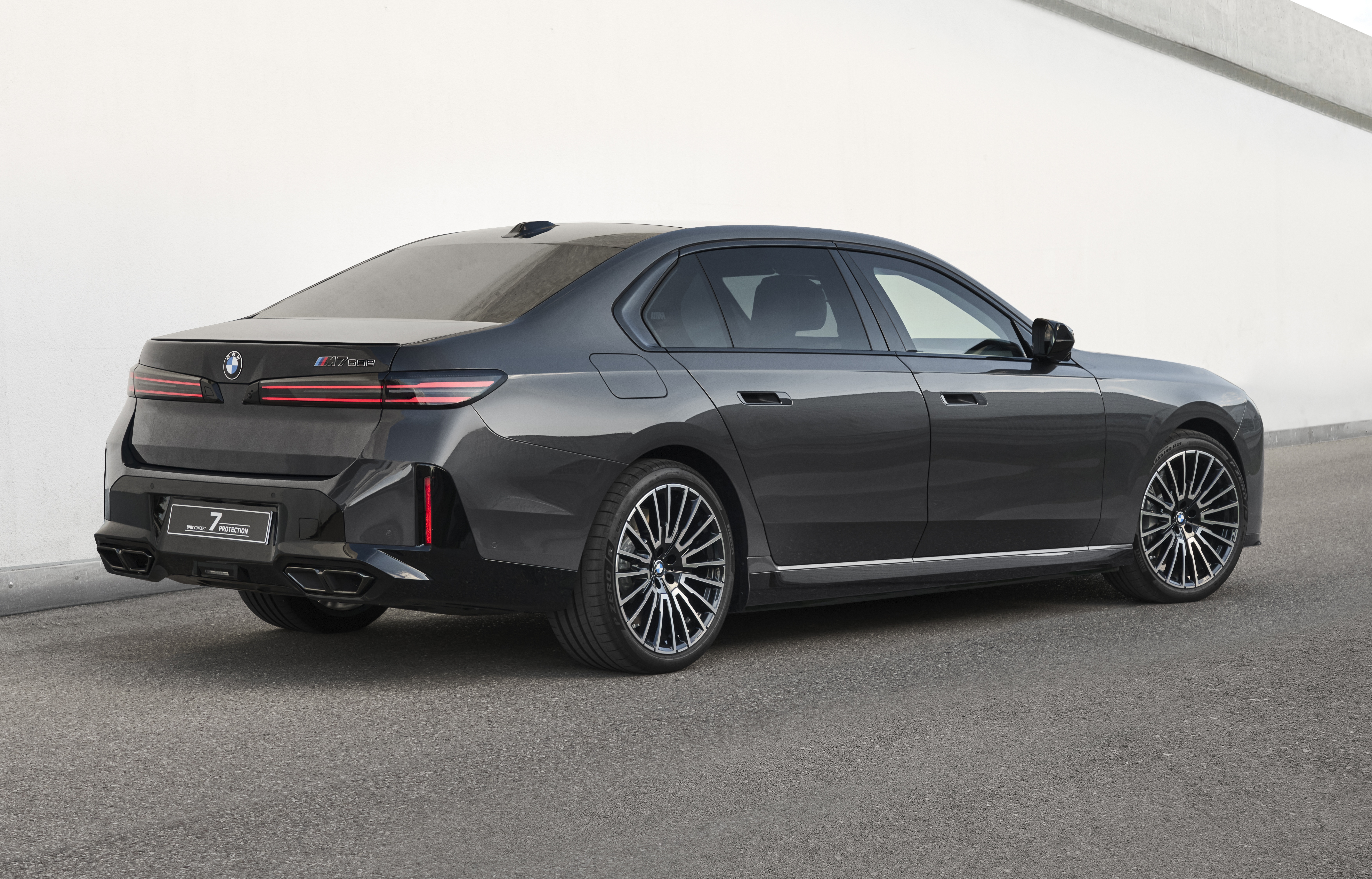 Sedã BMW M760e xDrive cinza escuro, visto de trás e lateral, estacionado em asfalto cinza com parede branca ao fundo. O carro tem rodas esportivas e quatro saídas de escape
