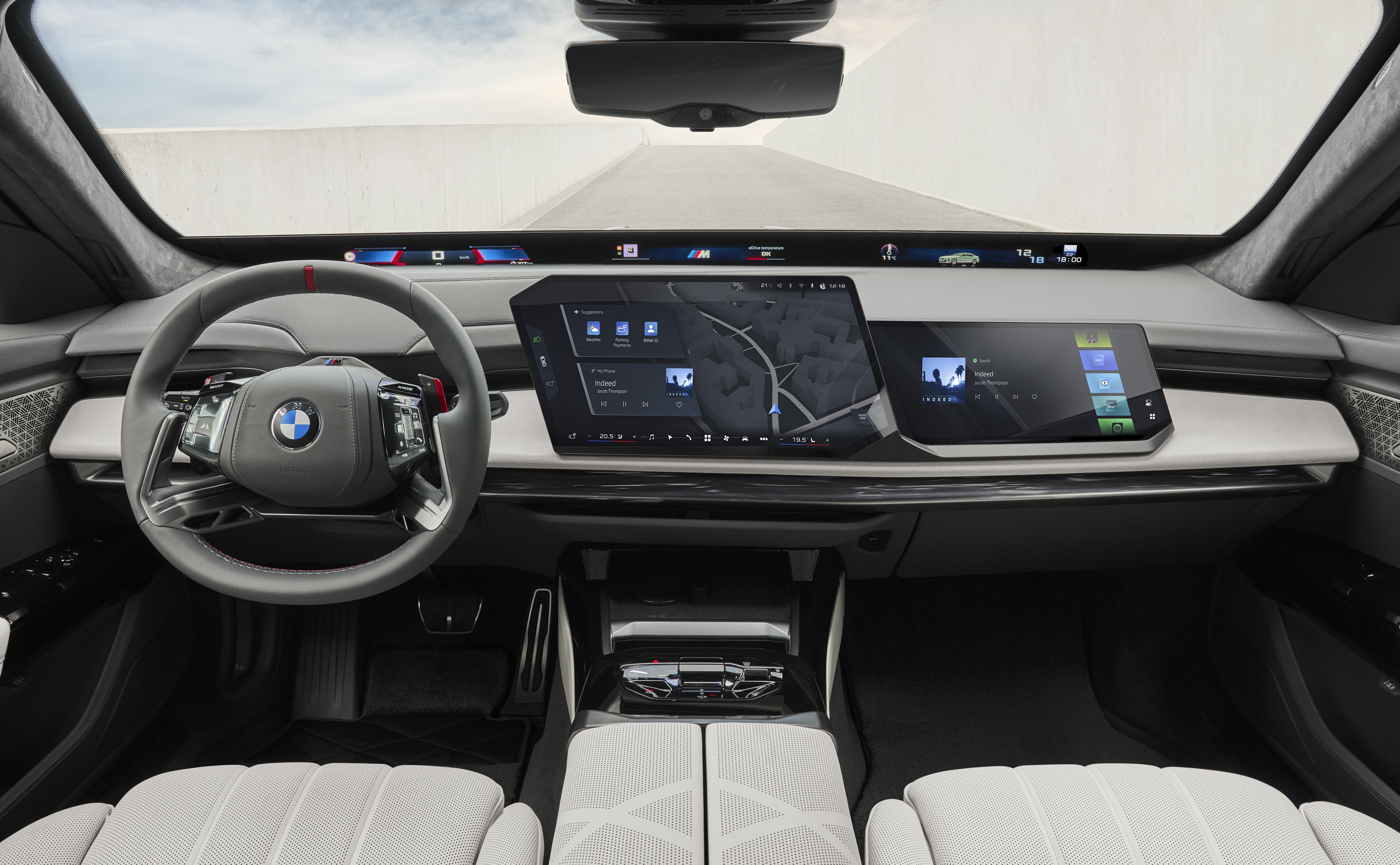 Interior de um carro moderno com volante cinza e logo BMW, painel digital curvo exibindo mapa e ícones, e bancos dianteiros claros com textura geométrica. O para-brisa mostra uma estrada cinza e céu claro
