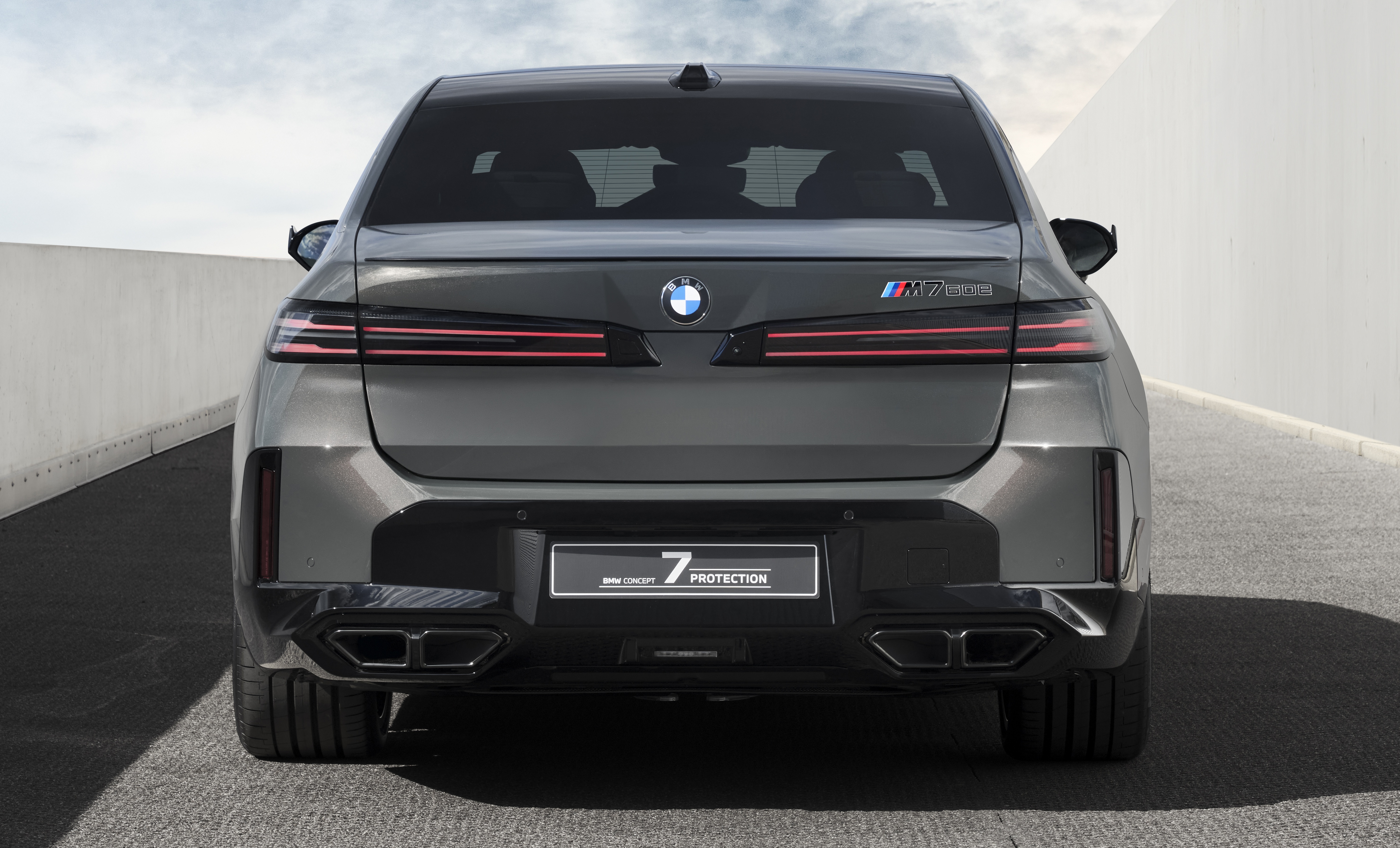 Traseira de um carro BMW cinza escuro, modelo 7 Protection, estacionado em uma rampa de concreto. O veículo possui lanternas horizontais finas com luzes vermelhas acesas, emblema BMW centralizado, e na lateral direita, o logo M760e. Abaixo, a placa preta exibe BMW CONCEPT 7 PROTECTION. O para-choque traseiro é preto com duas saídas de escapamento retangulares em cada lado. O céu está nublado