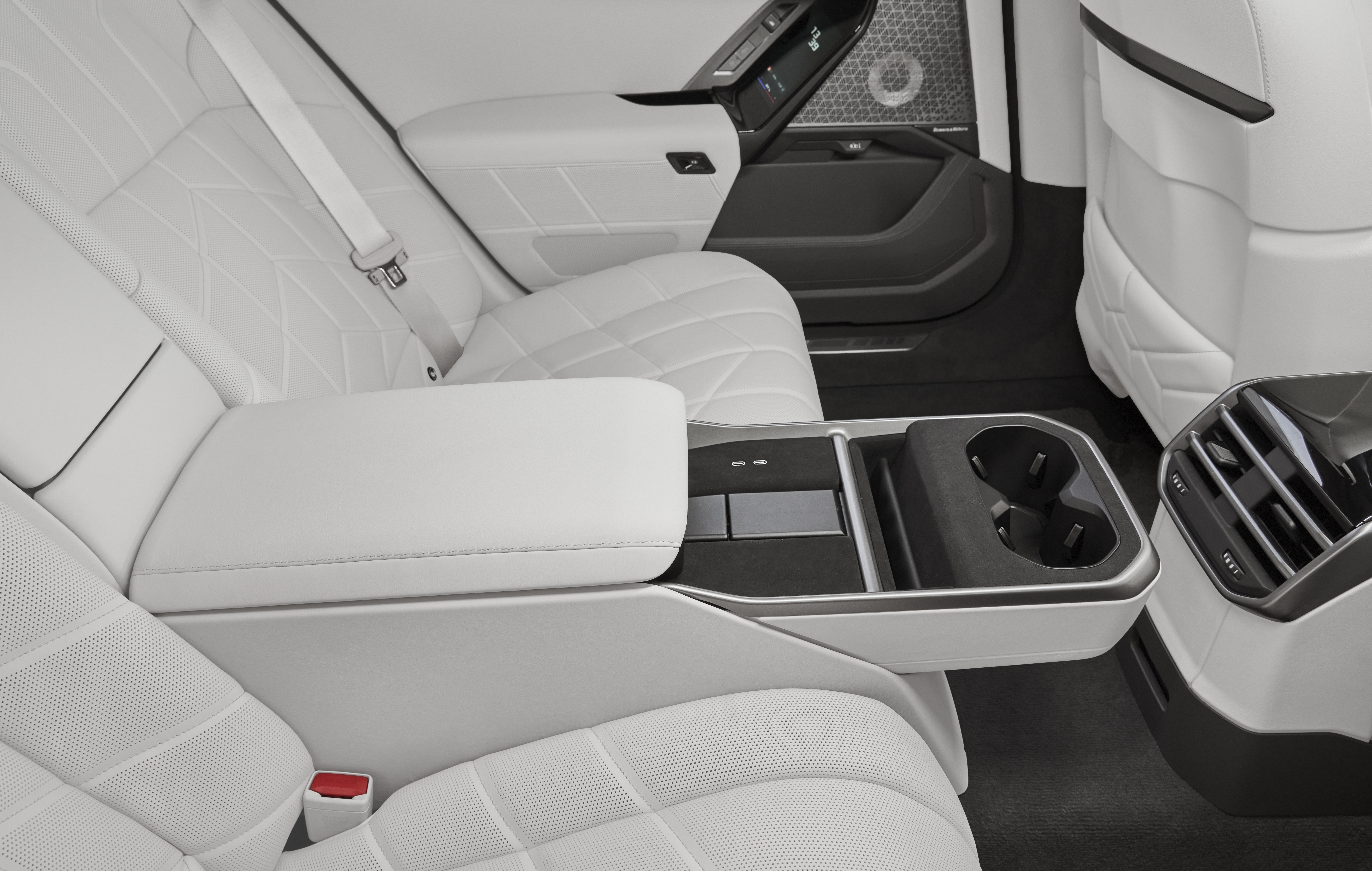 Interior de carro de luxo com bancos traseiros brancos acolchoados, cinto de segurança e apoio de braço central rebatível. O apoio revela dois porta-copos e compartimentos de armazenamento. Ao fundo, a porta e parte do painel com tela digital e alto-falante.