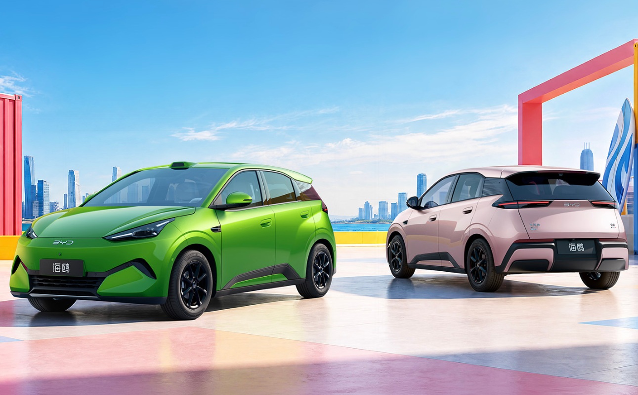 Dois carros elétricos compactos, um verde e outro rosa, estacionados em uma área colorida com um contêiner vermelho e uma prancha de surfe, com uma cidade costeira ao fundo sob céu azul