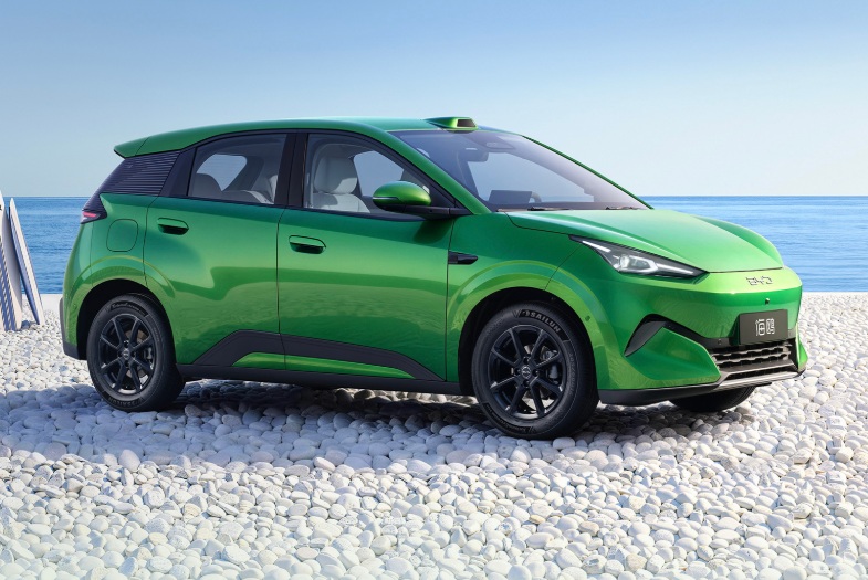 Carro elétrico verde-limão, modelo hatchback, estacionado em uma praia de pedras brancas, com o oceano azul ao fundo e pranchas de surfe à esquerda