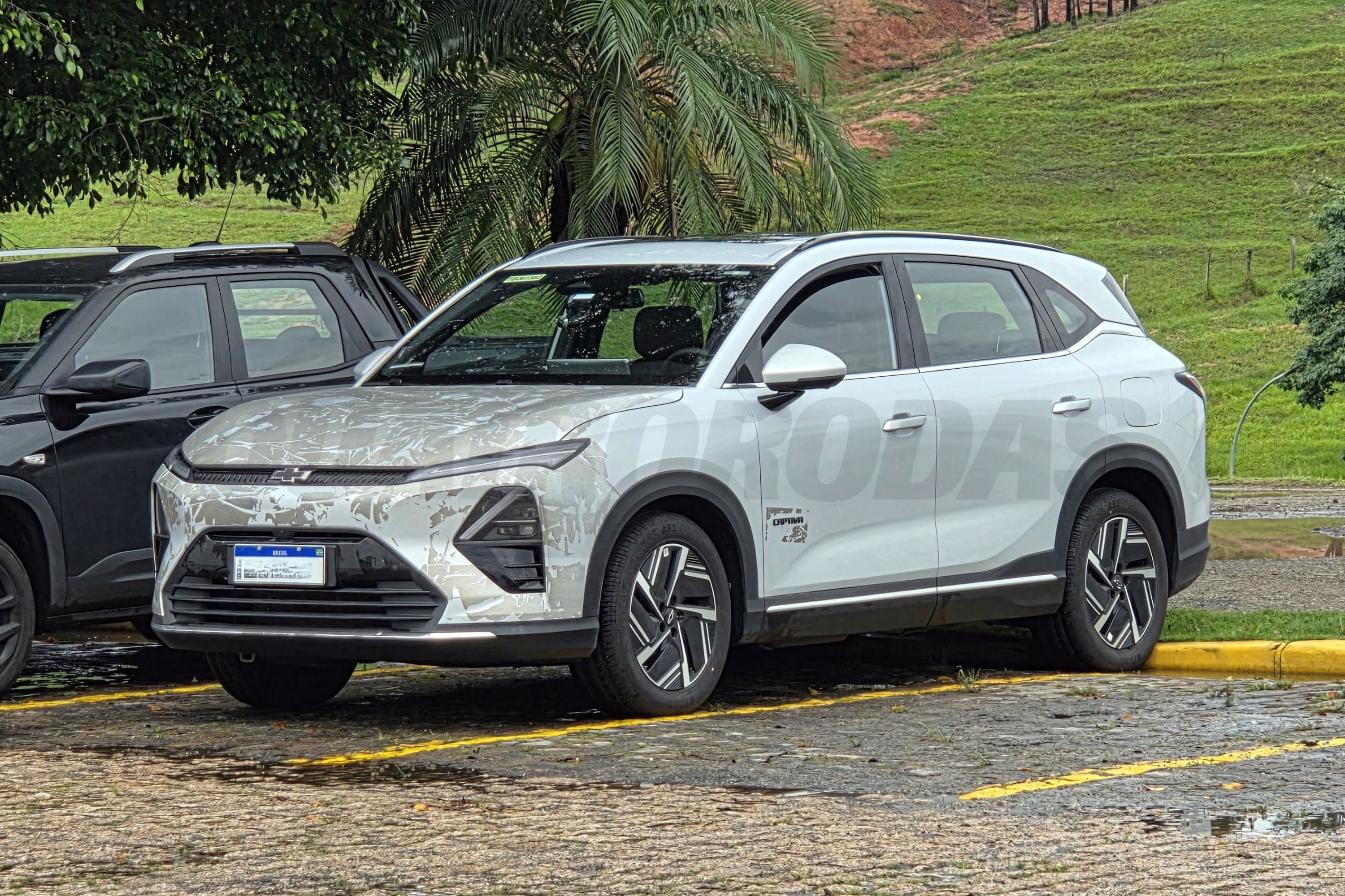 SUV branco camuflado com adesivos, estacionado em uma vaga de paralelepípedos com faixas amarelas, ao lado de uma picape preta. Ao fundo, vegetação verde e um morro.