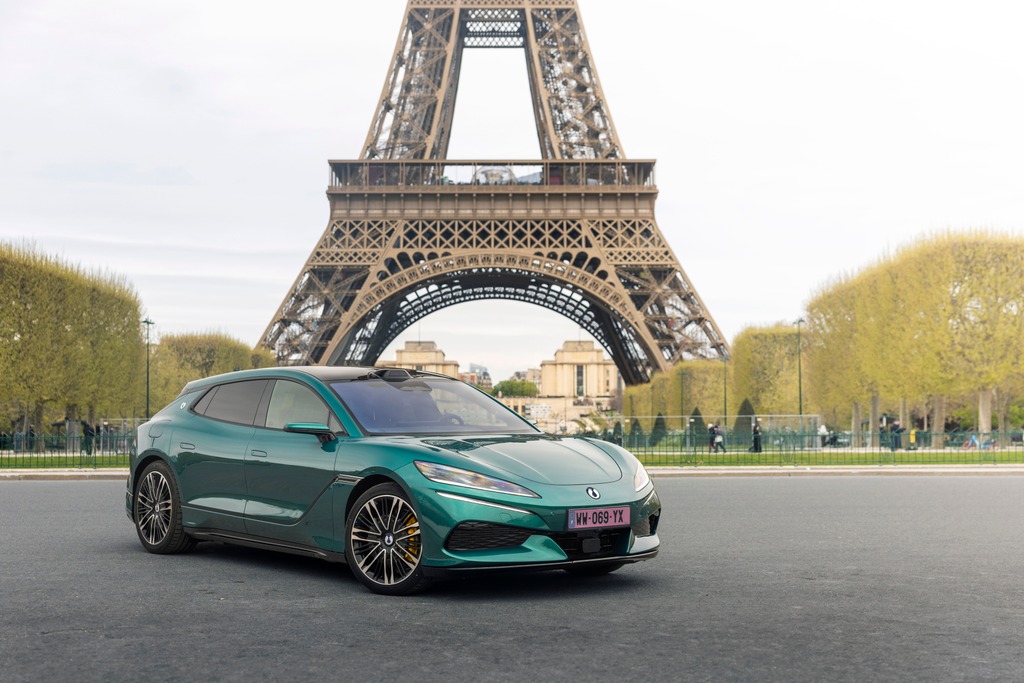 Carro elétrico verde-azulado, modelo station wagon, estacionado em rua pavimentada com a Torre Eiffel ao fundo, em Paris. O veículo tem rodas esportivas e placa rosa