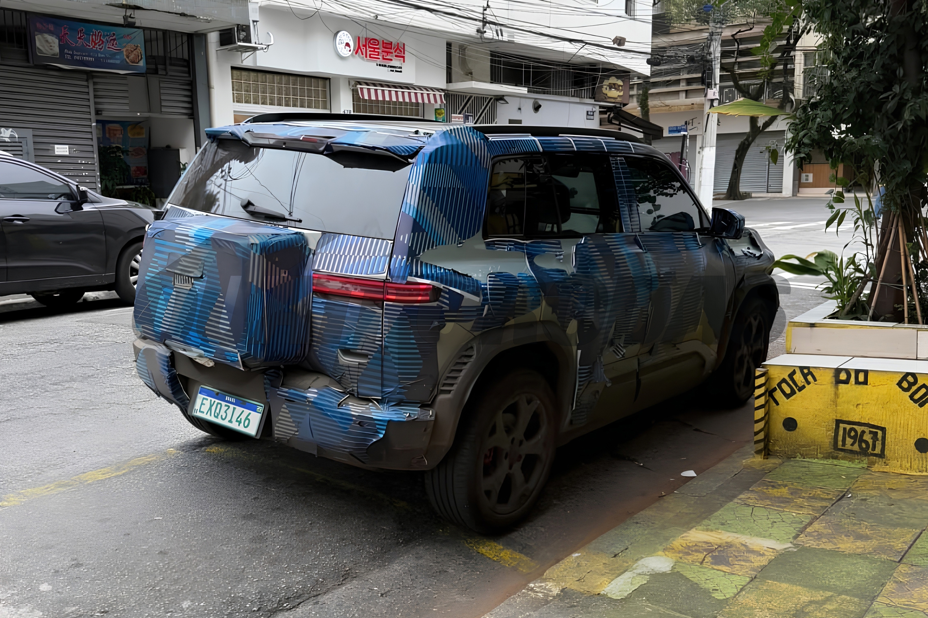 SUV camuflado com adesivos azuis e pretos, estacionado na rua ao lado de um canteiro de plantas e um bloco de concreto amarelo com a inscrição TOCA DO BOI 1967. Ao fundo, prédios comerciais e outro carro escuro