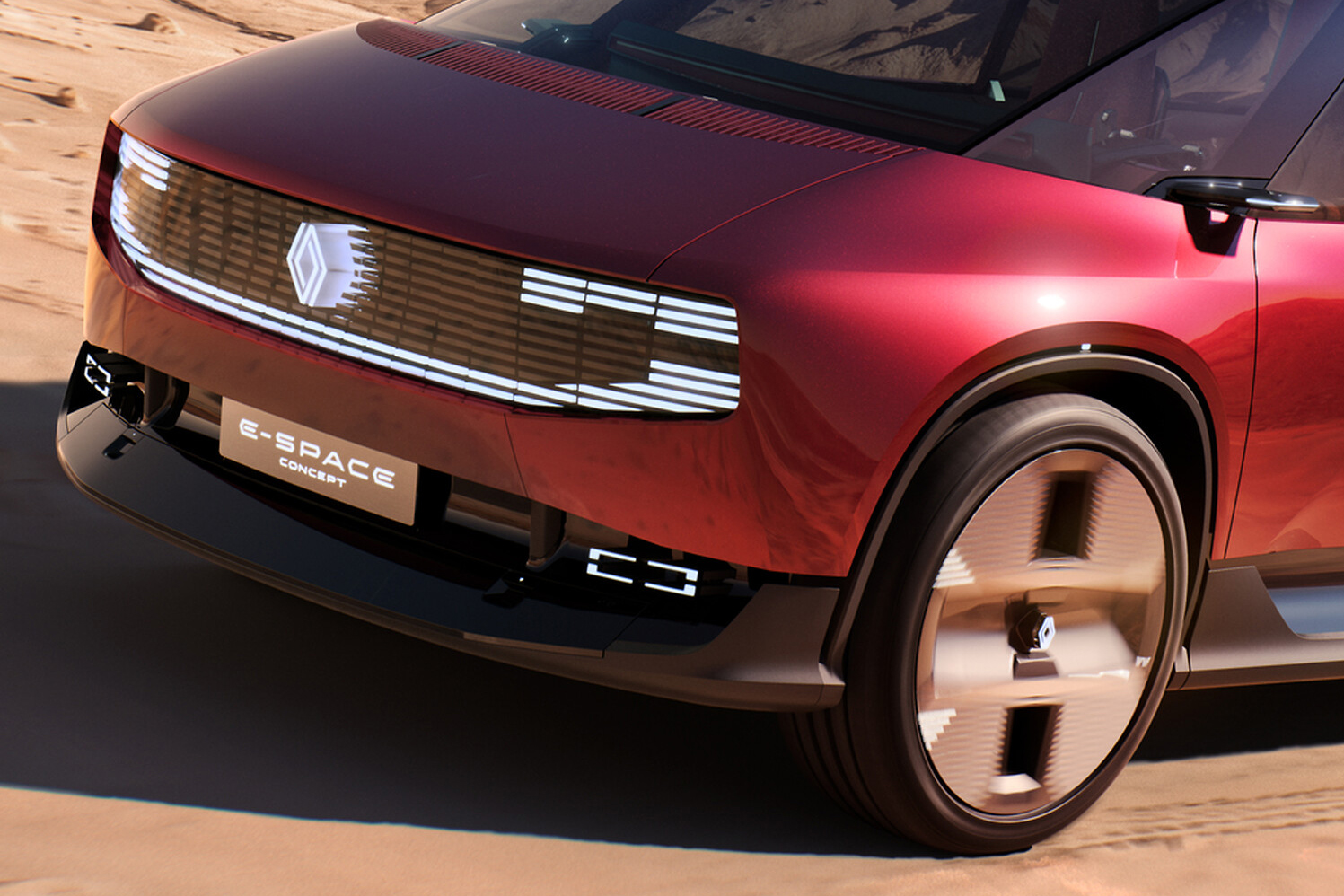 Parte frontal de um carro conceito Renault E-Space vermelho-cereja, com grade iluminada por LEDs brancos e o logotipo da marca no centro. A placa E-SPACE CONCEPT está no para-choque preto, e a roda dianteira direita tem um design futurista com raios largos e cor bronze, em um cenário desértico