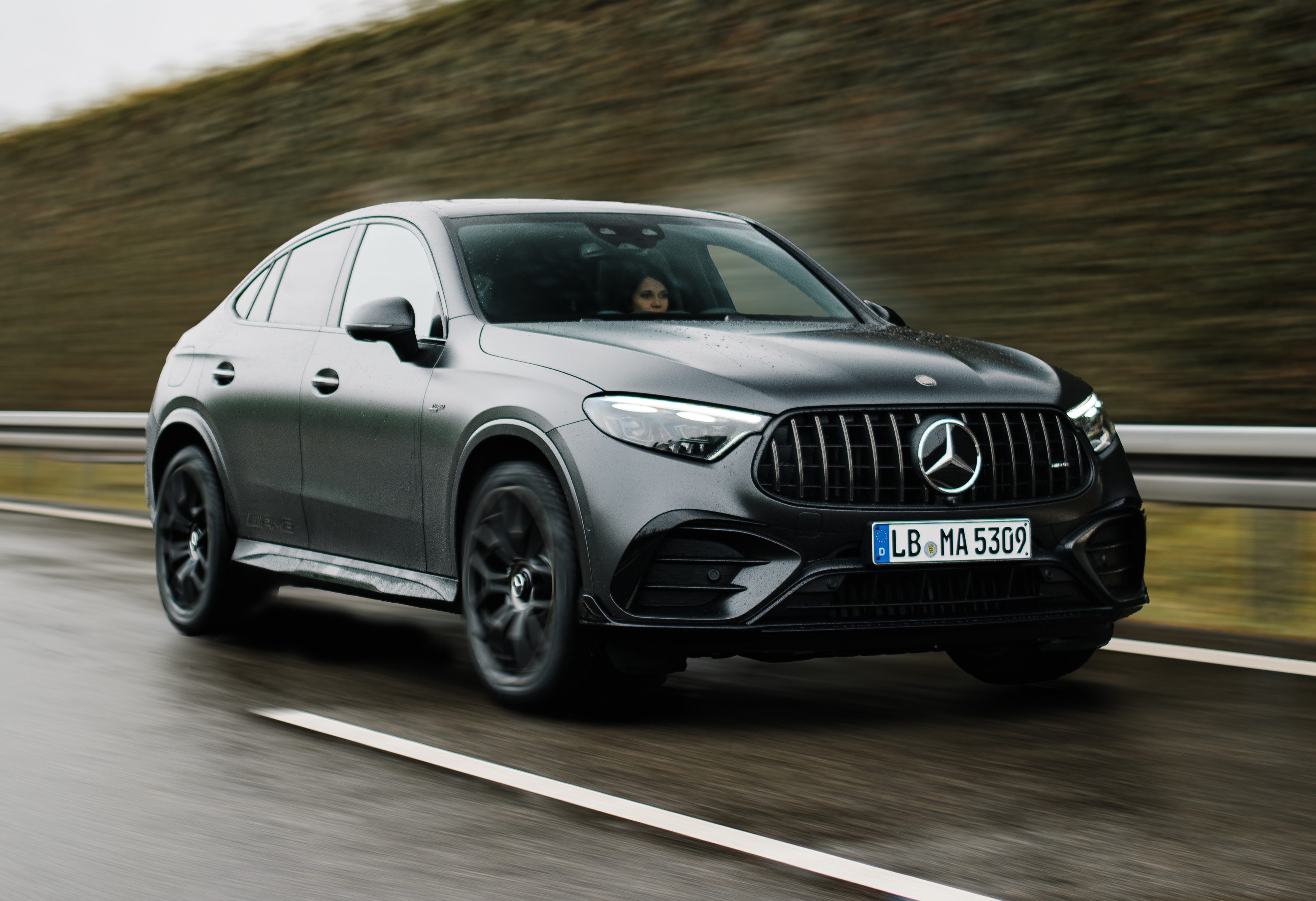 SUV cupê Mercedes-Benz GLC 53 AMG preto fosco em movimento na estrada molhada, com uma pessoa ao volante. O carro tem rodas pretas e grade frontal vertical com o logotipo da Mercedes. Ao fundo, um barranco coberto por vegetação marrom e o céu cinzento