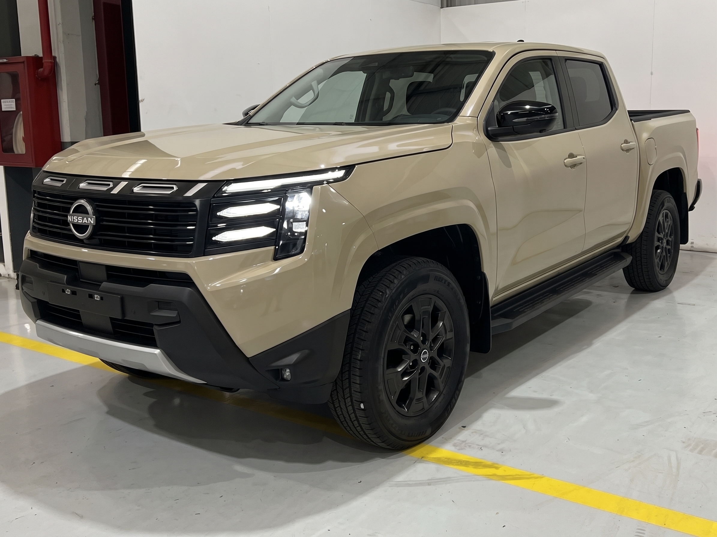 Picape Nissan Frontier Pro-4X 2023 bege, vista de frente e lateral, com faróis acesos e rodas pretas, estacionada em piso cinza com faixa amarela