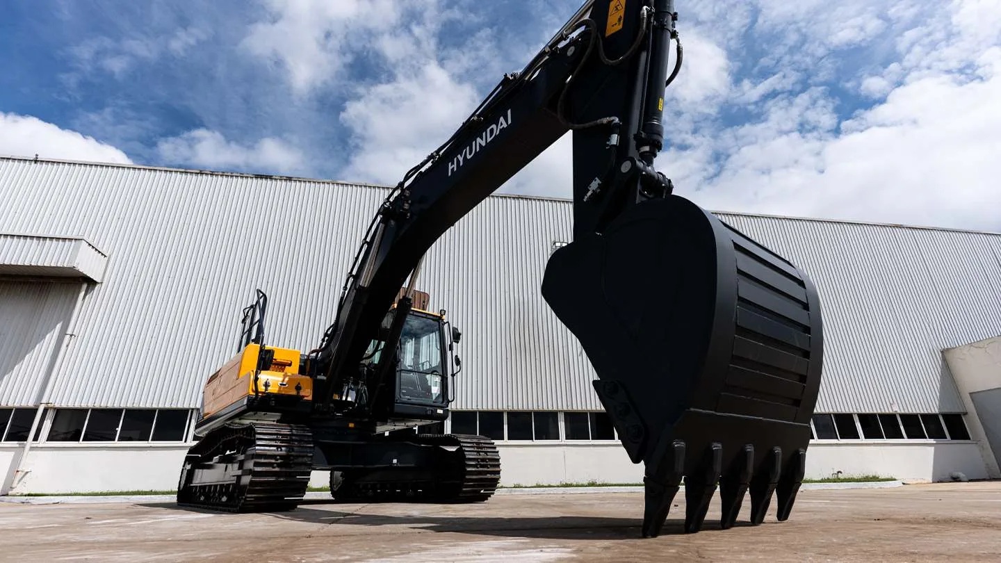 Escavadeira Hyundai preta e amarela com esteiras, braço estendido e caçamba grande em primeiro plano, estacionada em pátio de concreto com um galpão branco ao fundo sob céu azul com nuvens