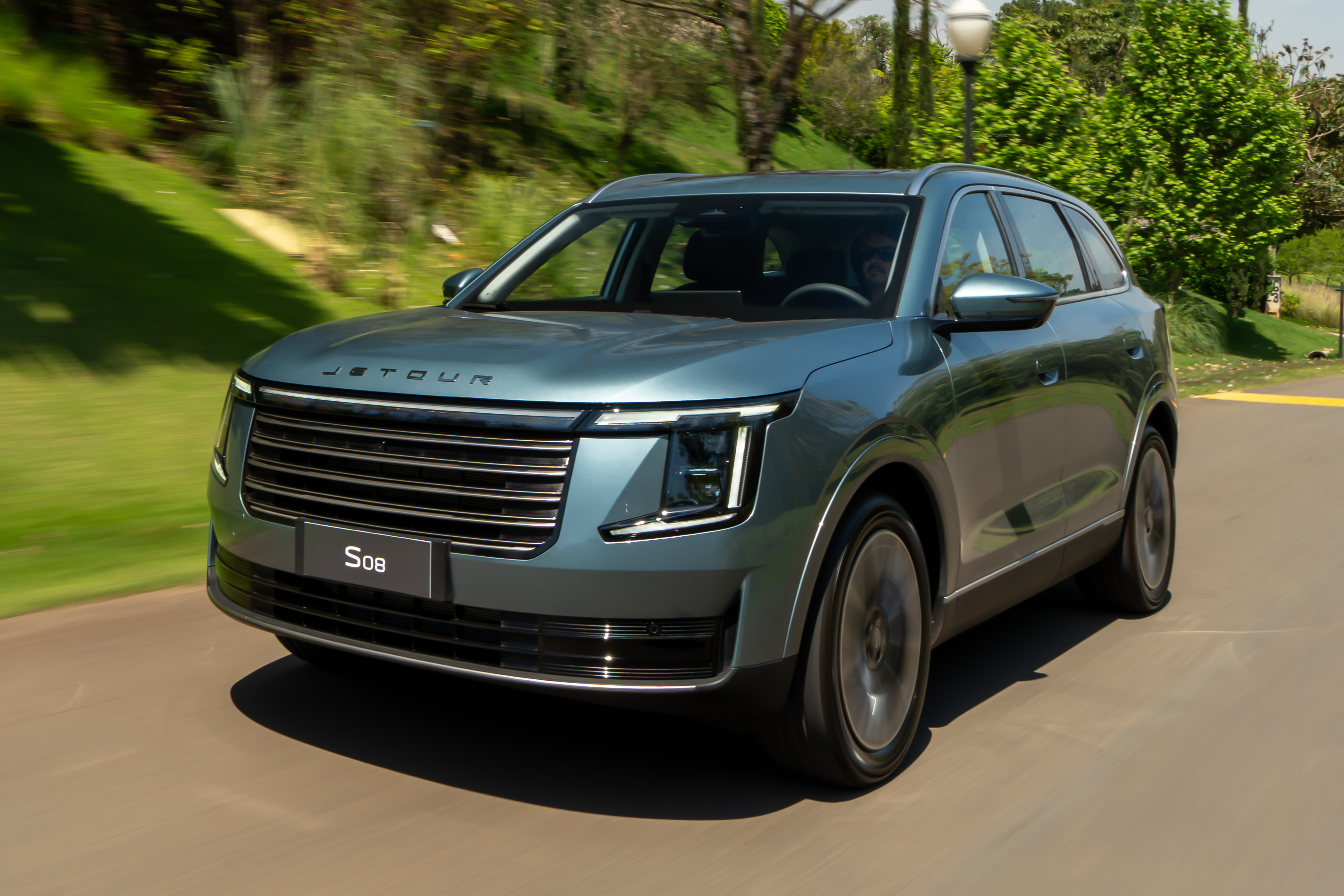 SUV Jetour S08 azul-esverdeado em movimento, visto de frente e levemente lateral, em uma estrada asfaltada com vegetação verde e um poste de luz ao fundo