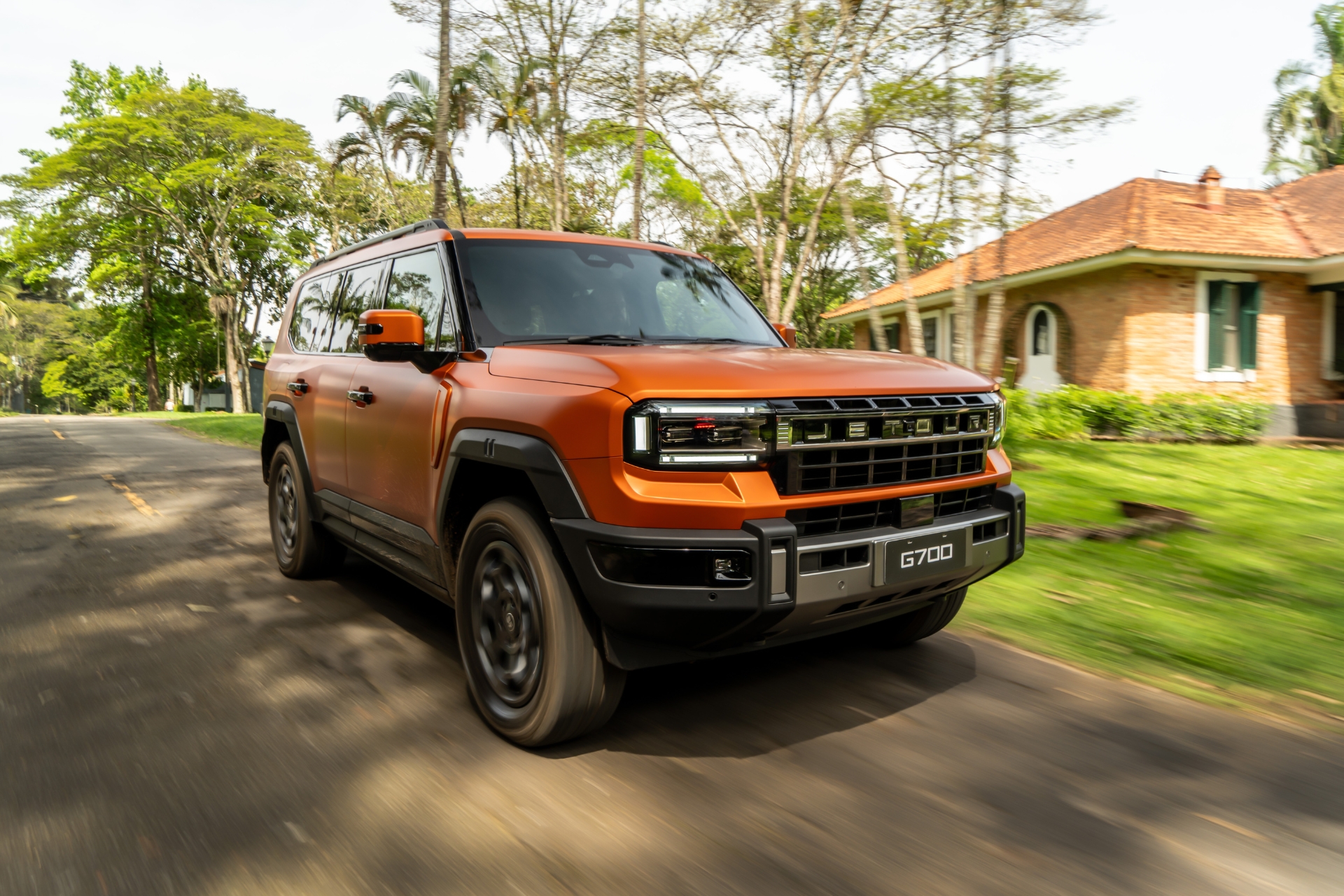 SUV laranja fosco, modelo G700, em movimento numa estrada asfaltada com faixa amarela. Ao fundo, árvores verdes e uma casa de tijolos com telhado de cerâmica.