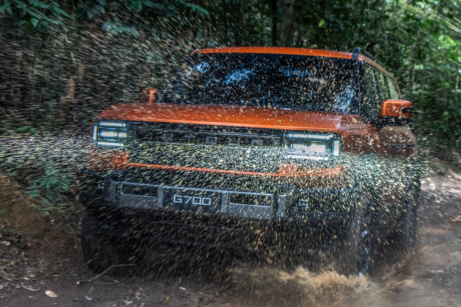 SUV laranja G700 em movimento, levantando muita água e lama em uma trilha de terra com vegetação densa ao fundo