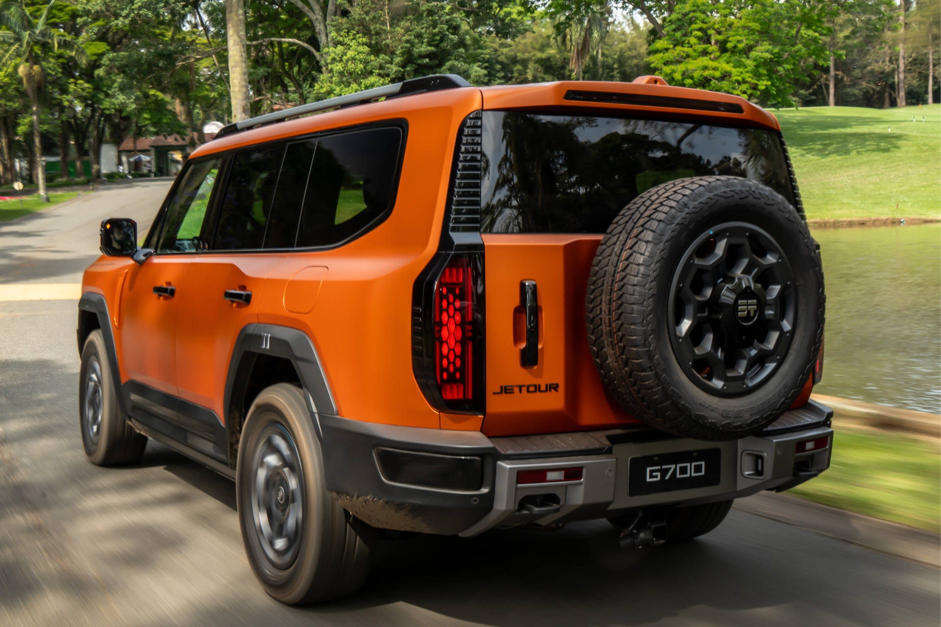 SUV Jetour G700 laranja, visto de traseira lateral, com estepe na tampa do porta-malas, rodando em estrada asfaltada. Ao fundo, árvores e um lago.