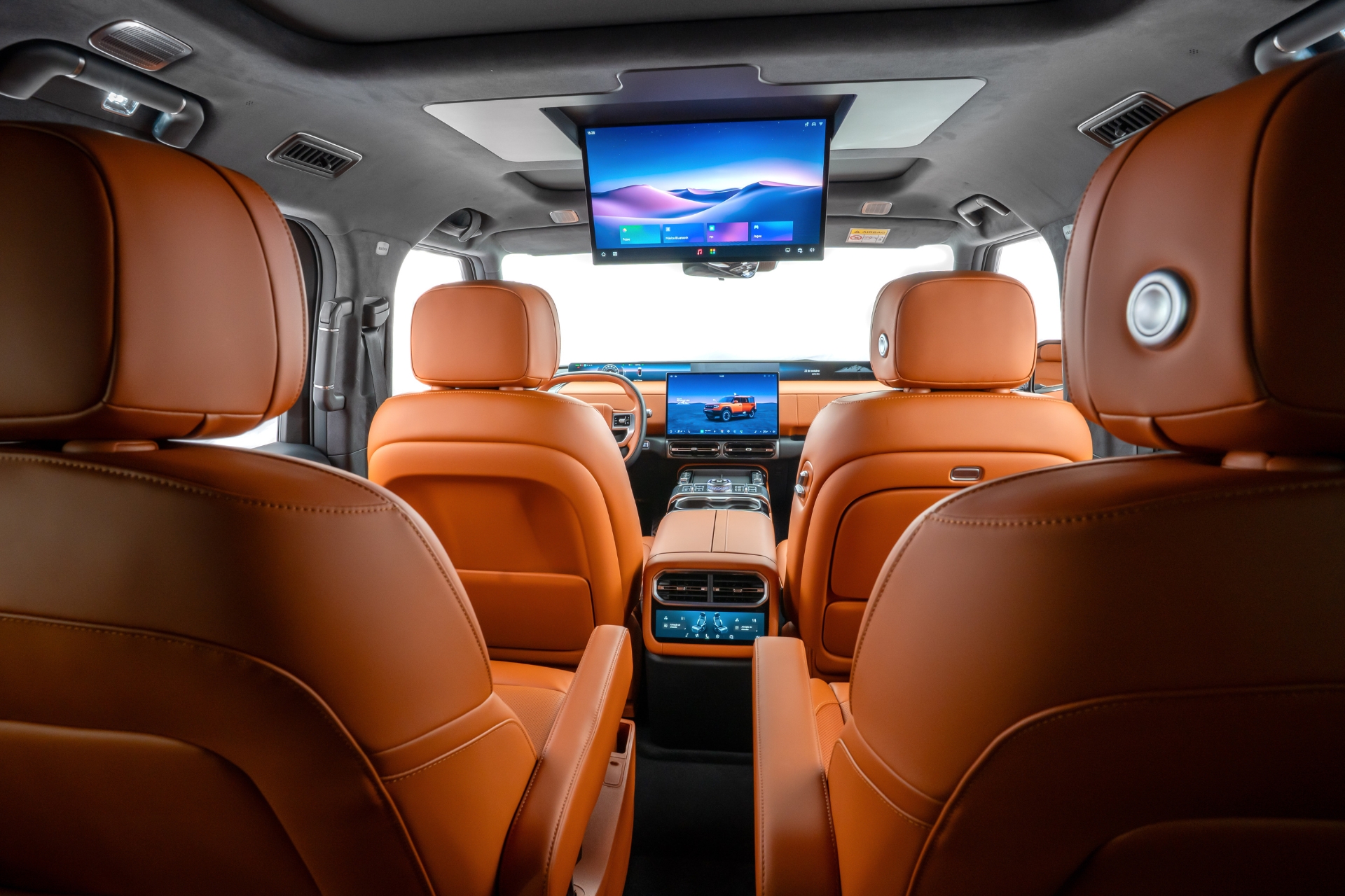 Interior luxuoso de um carro com bancos de couro marrom-claro, vistos da segunda fila. Há uma tela grande no teto exibindo uma paisagem montanhosa e outra menor no painel frontal com um carro laranja.