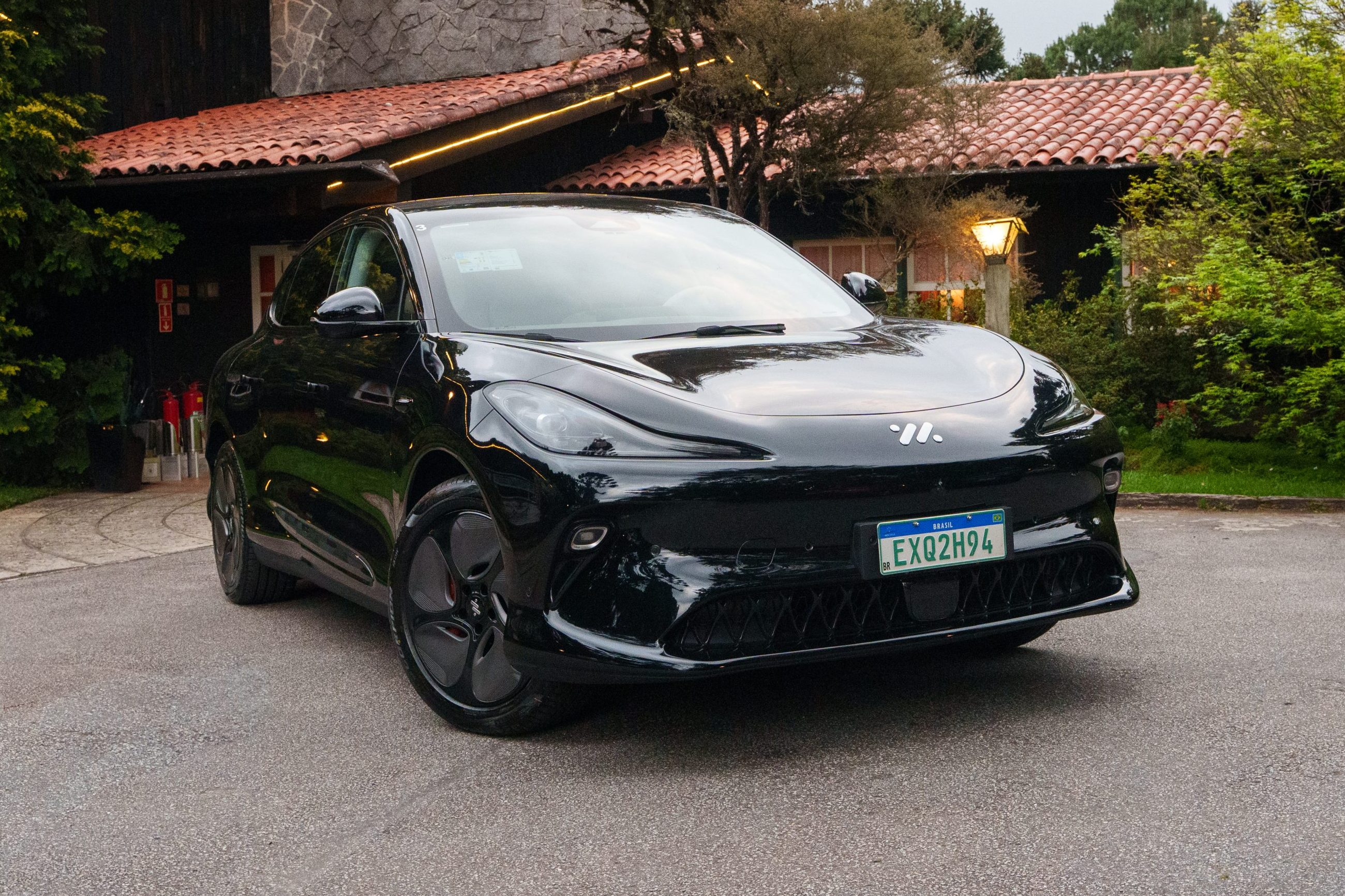 Carro elétrico preto, modelo Smart #1, estacionado em frente a uma casa com telhado de telhas e detalhes em pedra, cercada por árvores e arbustos verdes, sob um céu nublado