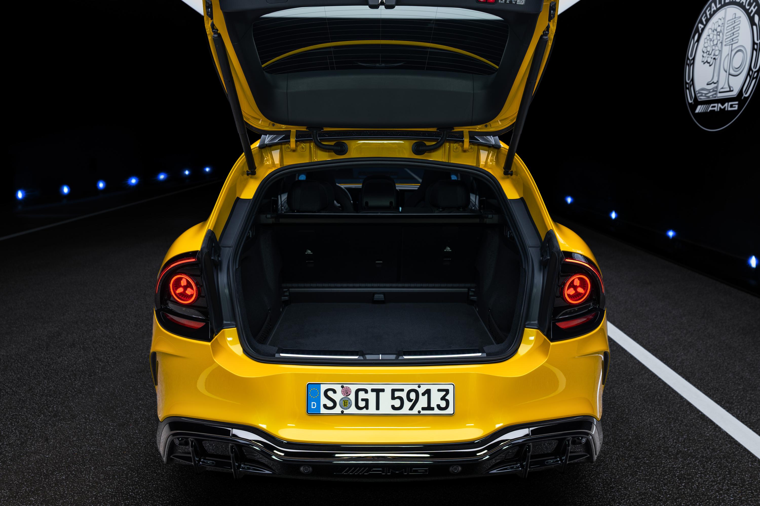 Traseira de um carro esportivo amarelo com o porta-malas aberto, revelando o interior escuro e vazio. A placa de licença exibe S GT 5913. As lanternas traseiras vermelhas estão acesas, e o veículo está em uma estrada escura com luzes azuis no horizonte e uma faixa branca à direita. O logo AMG aparece no canto superior direito