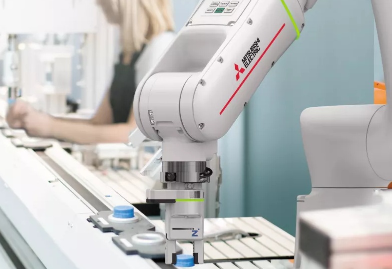 Braço robótico branco da Mitsubishi Electric com detalhes vermelhos e verdes, manipulando uma peça com tampa azul em uma linha de montagem. Ao fundo, uma pessoa loira desfocada trabalha em outra estação, sugerindo automação industrial e colaboração entre humanos e robôs