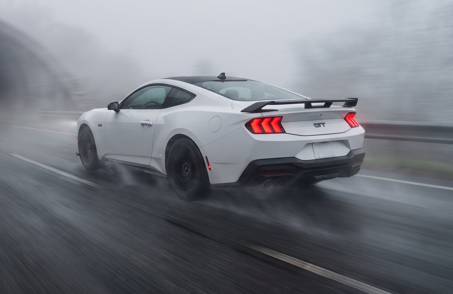 Carro esportivo branco, modelo Mustang GT, visto de traseira lateral, acelerando em estrada molhada sob neblina. As lanternas traseiras vermelhas estão acesas, e há um aerofólio preto na tampa do porta-malas.