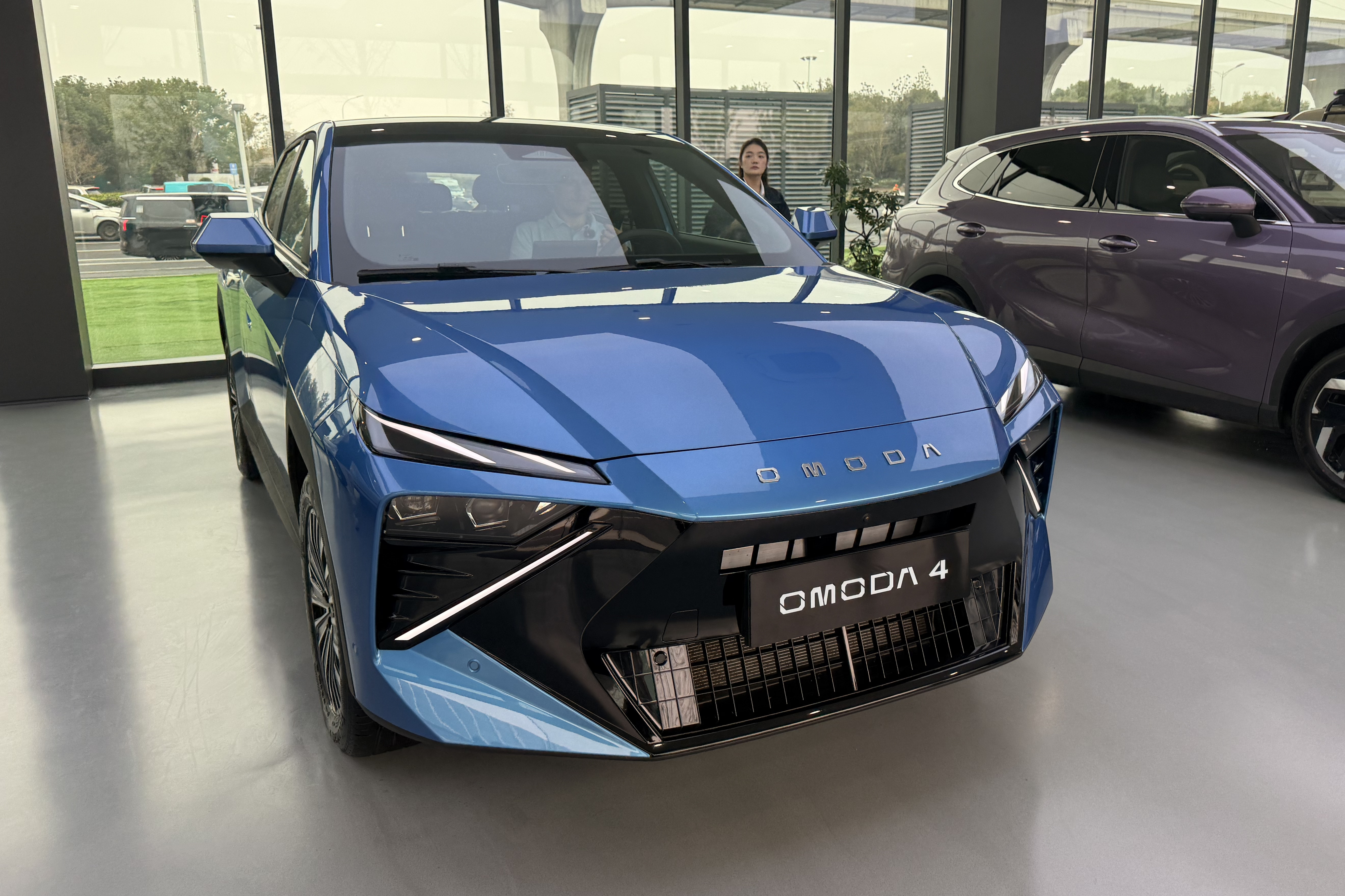Carro OMODA 4 azul metálico em destaque no centro, com design moderno e faróis afilados. Uma mulher asiática de blusa escura observa ao fundo, perto de uma janela que reflete árvores e outros veículos. Um SUV roxo está parcialmente visível à direita.